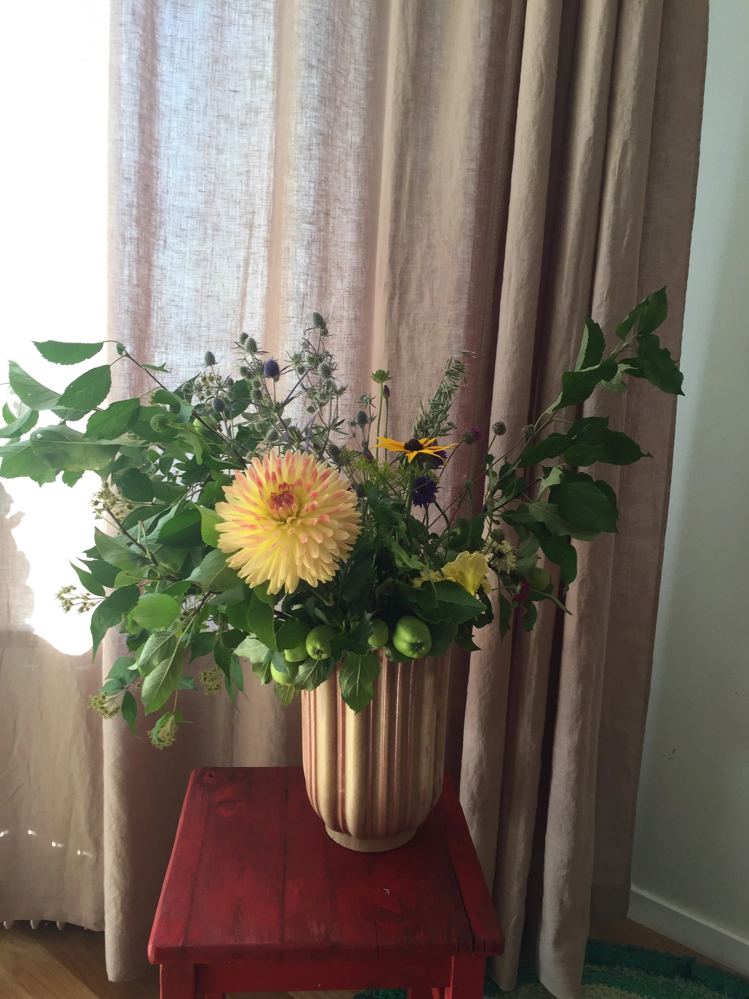 En blomsterbukett i en guldfärgad vas placerad på ett rött bord, med gardiner i bakgrunden. Dahlia, vildapel.