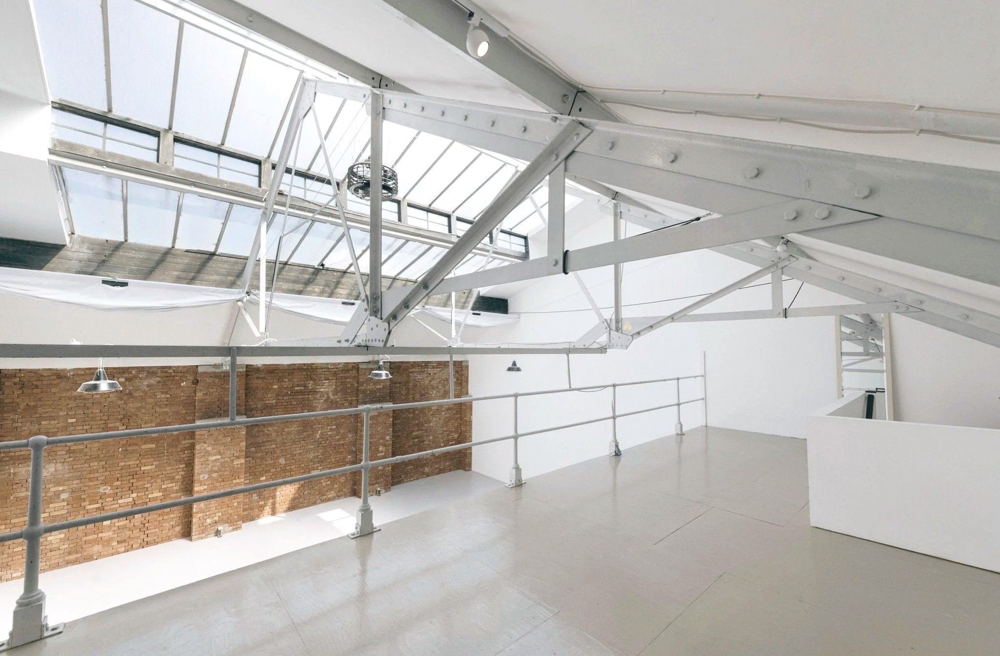 Interior of a modern building with white structural beams, a brick wall, a railing, and a ceiling with skylights.