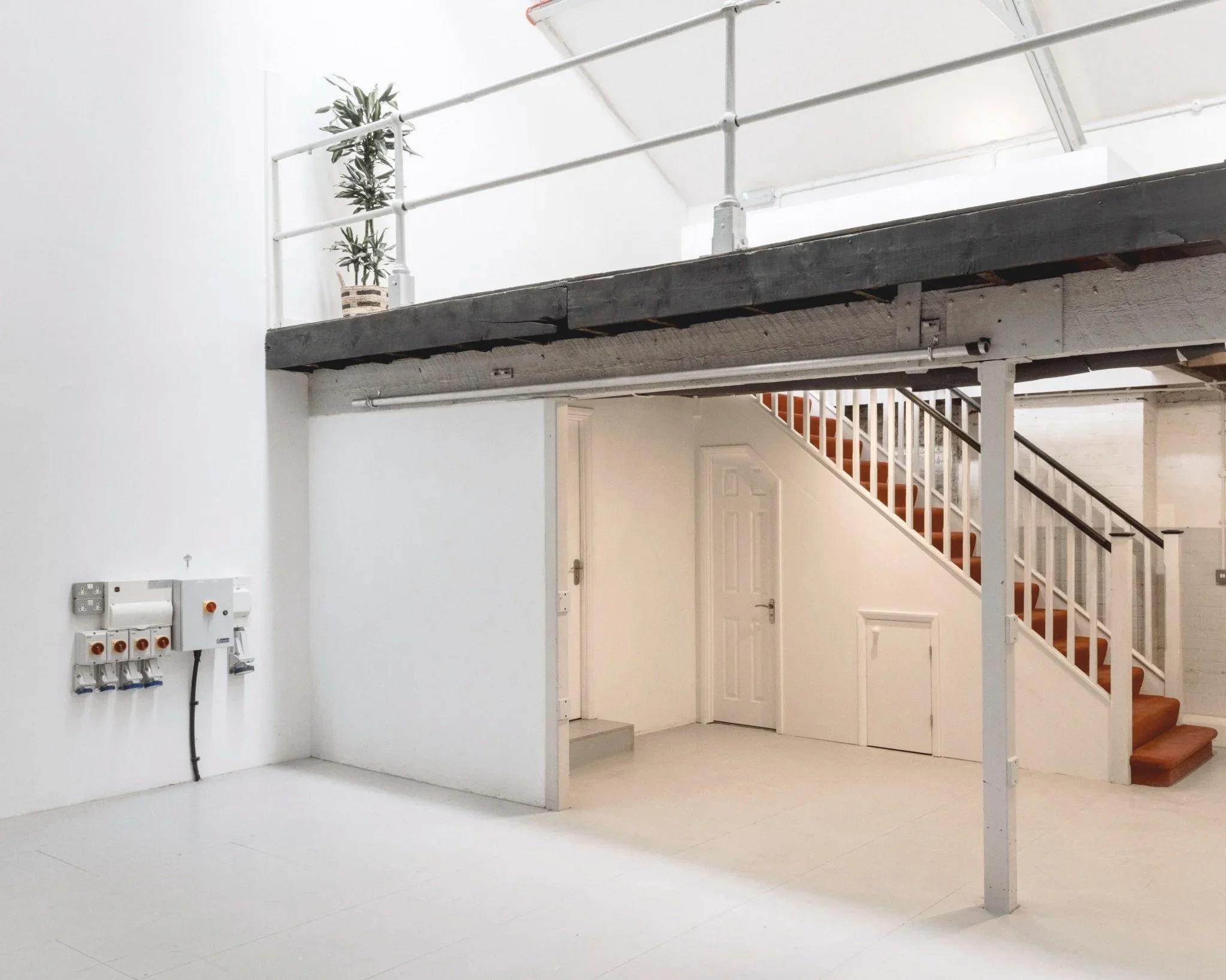 Interior of a minimalist, white-walled room with an upper loft area decorated with a potted plant, a staircase with wooden steps and a white railing, and an electrical panel on the wall.