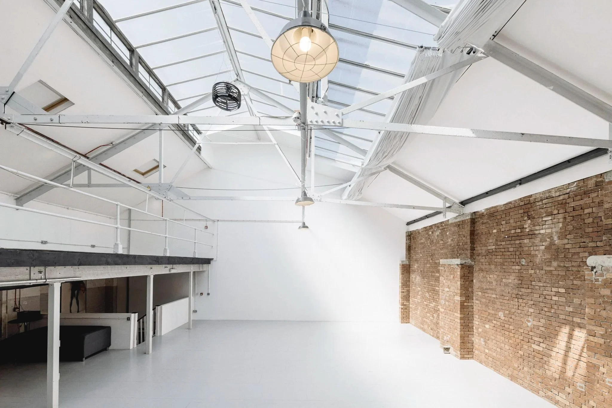 Interior of a spacious, modern industrial-style open-plan space with white walls, exposed brick wall on the right, a high ceiling with skylights, and hanging light fixtures.