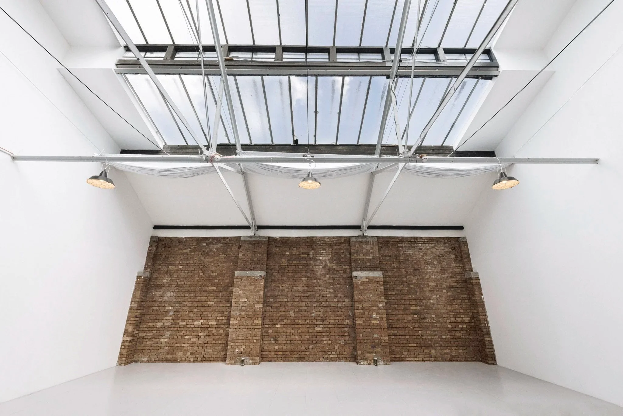 A room with a brick wall, white painted walls and ceiling, large skylight window with a grid of metal bars, and industrial-style hanging lights.