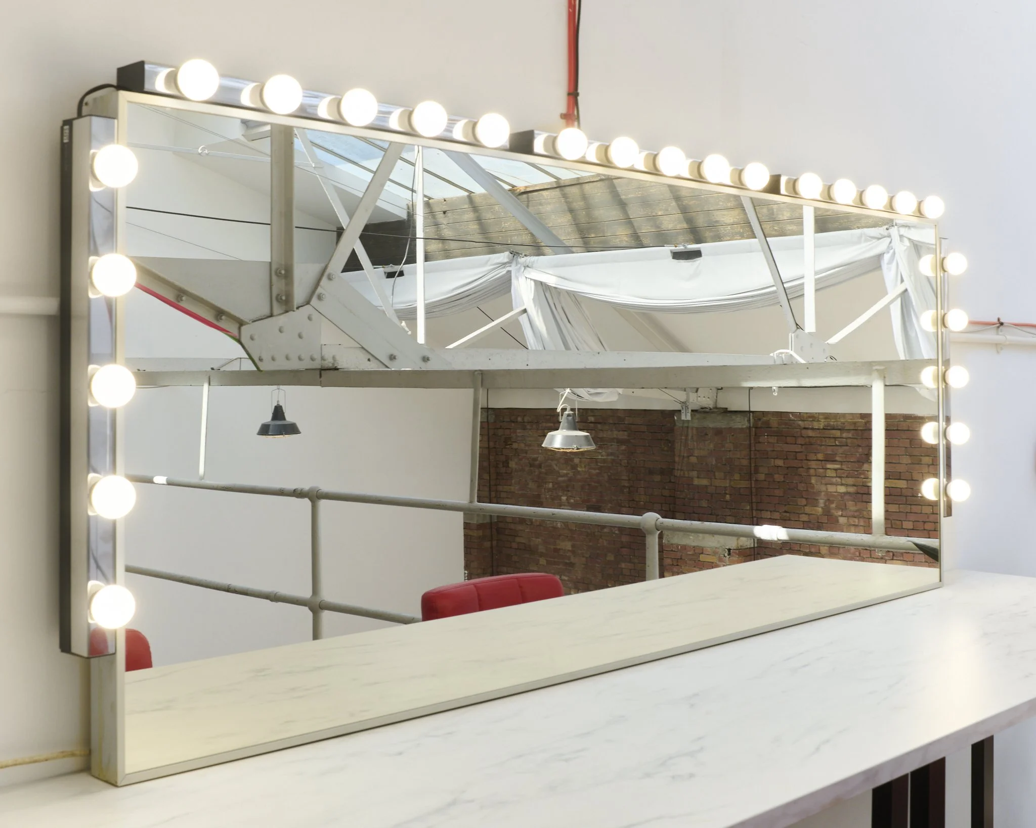 A large mirror with round vanity lights surrounding it, reflecting an industrial-style interior with brick walls, metal beams, and hanging lights.