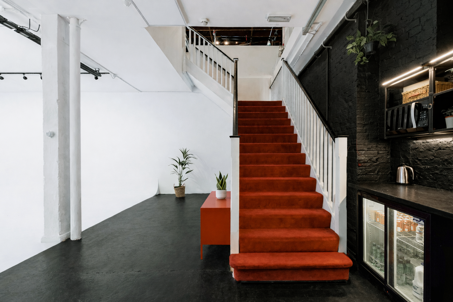Indoor space with a staircase covered in red carpet, white railings, and black walls. There is a small black table with a white potted plant, and a larger red table with a green plant in a basket. On the right side, lower shelves hold glass jars and 