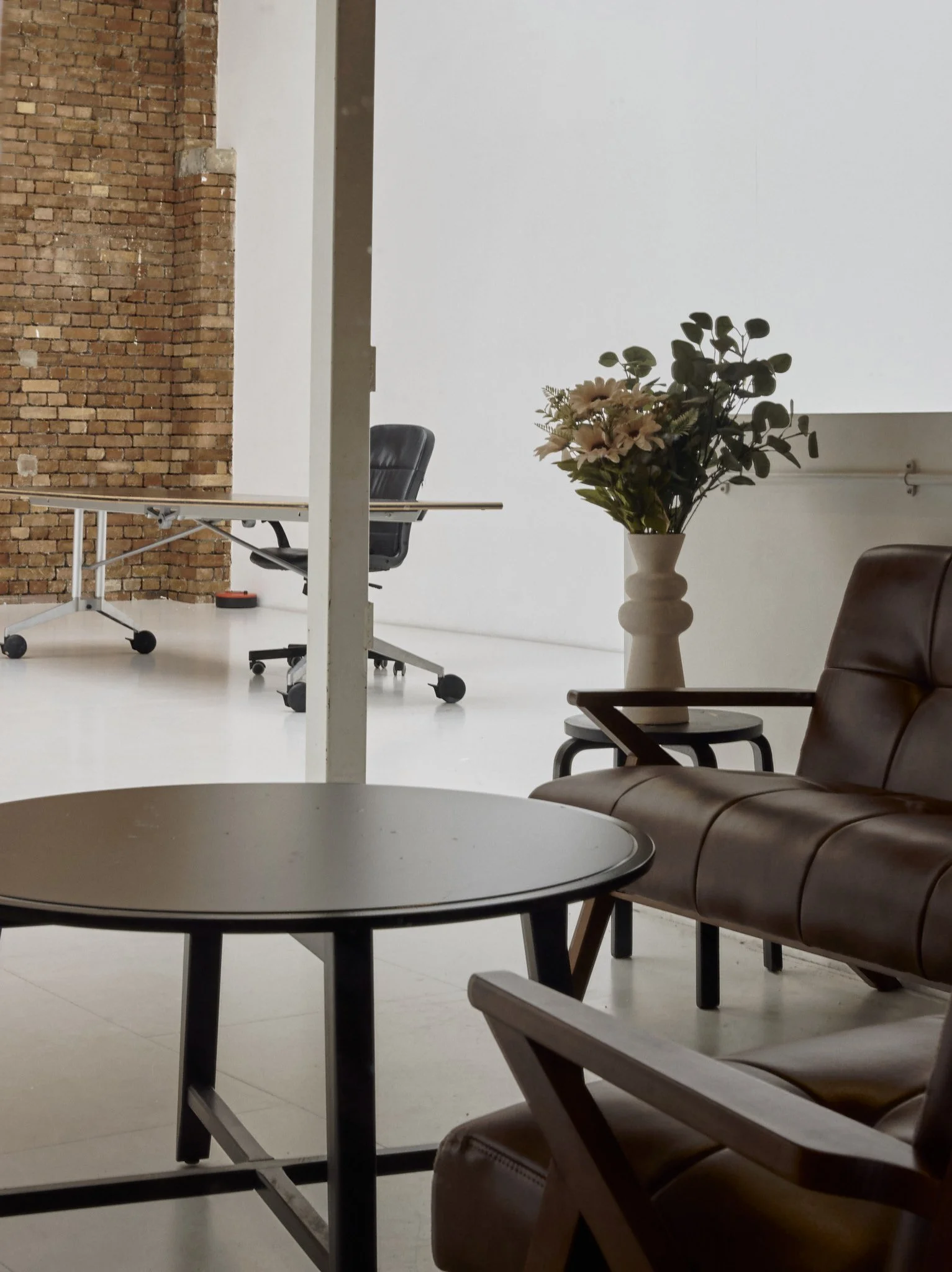 A cozy waiting area with brown leather chairs, a black round coffee table, a side table with a vase of flowers, and a workspace with a black office chair and table in the background, set against white walls and a brick wall.