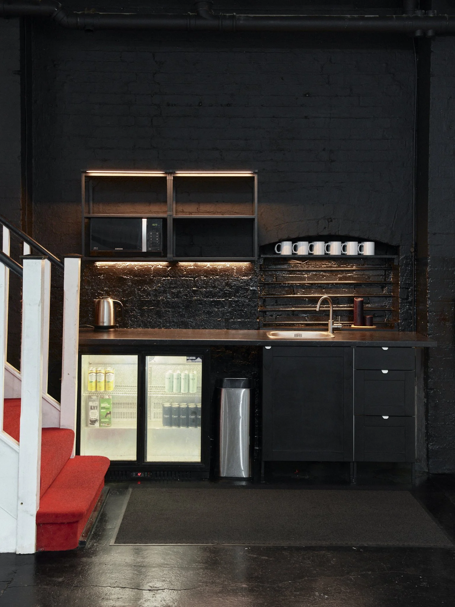 A small kitchen area with black brick wall, black cabinetry, stainless steel refrigerator, microwave, kettle, and a sink with a faucet. Seven white cups are on a shelf above the sink. A red carpeted stairway with white railings is on the left side of