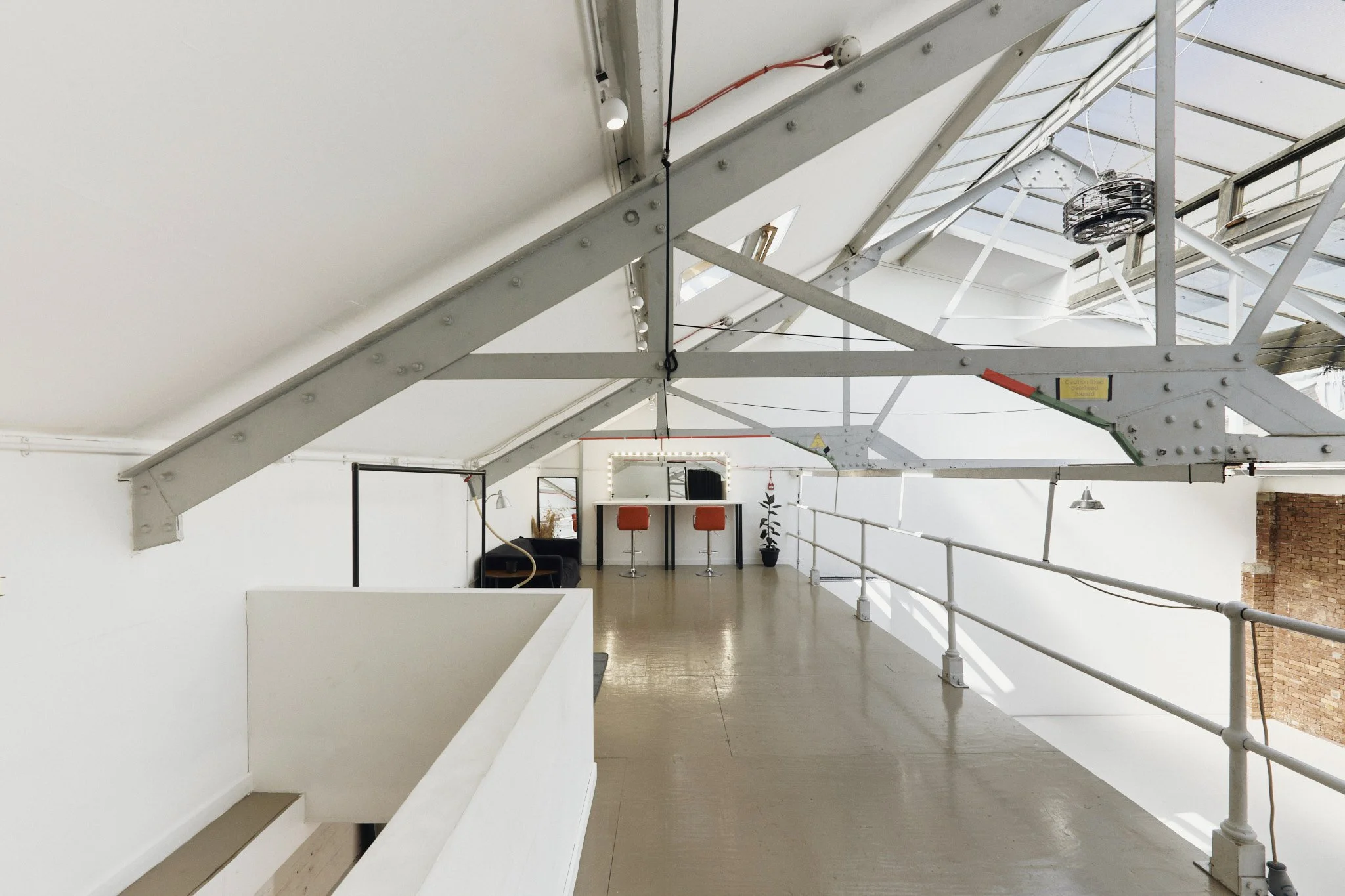 Interior of an industrial-style loft with a white ceiling and exposed metal beams, white walls, a small seating area with black couch and a bar table with two orange stools, and a potted plant.