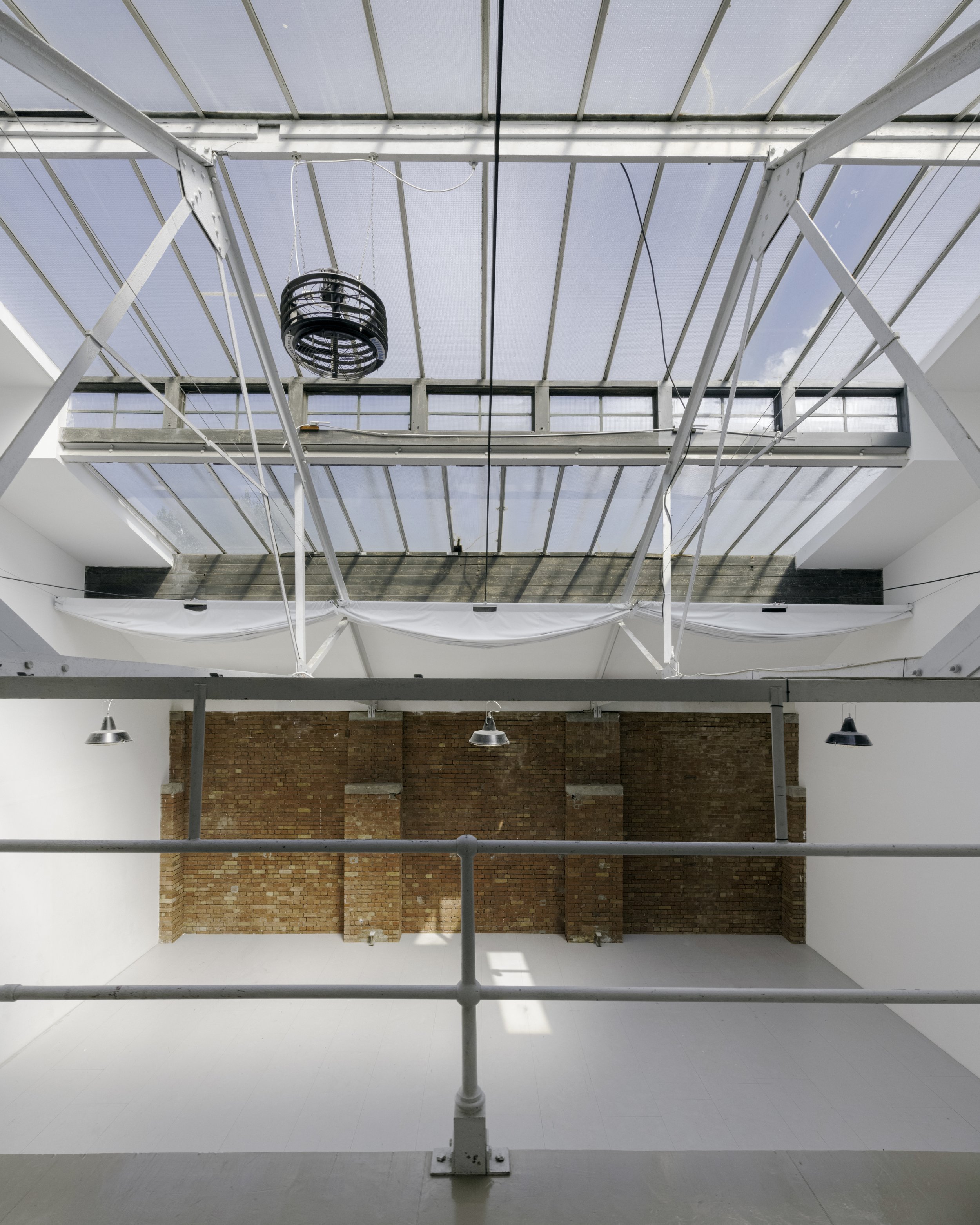 Interior view of a building with a large glass ceiling, exposed brick wall, and industrial lighting fixtures, taken from a mezzanine level with railings.