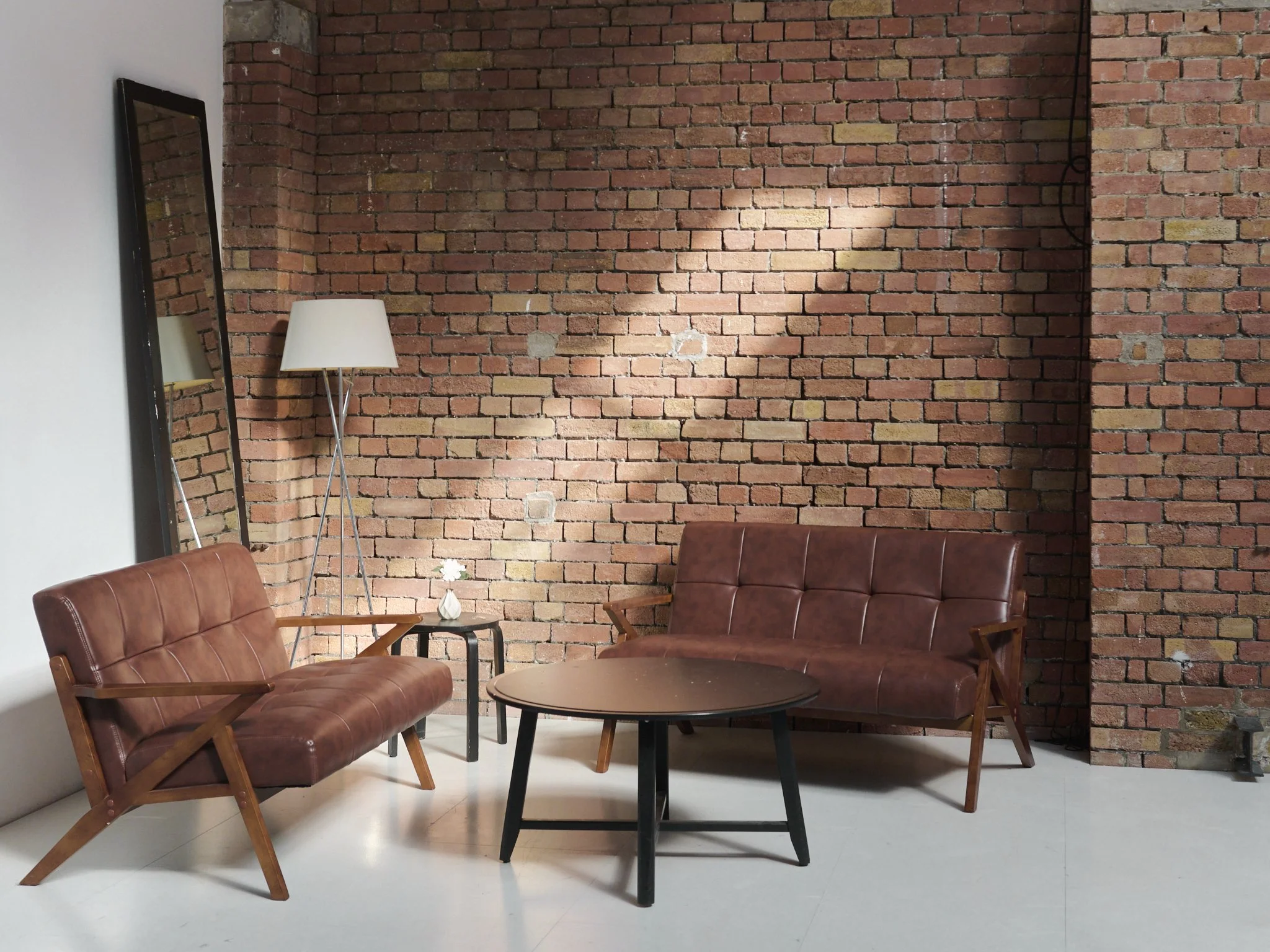 Living room with exposed brick wall, two brown leather sofas, a round black coffee table, a small side table with a white flower vase, a standing lamp, and a large rectangular mirror leaning against the wall.