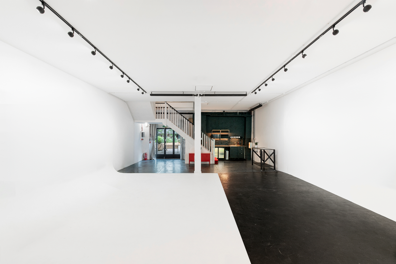 Empty art gallery with white walls, black floor, and track lighting on ceiling, with a small kitchenette area and stairs leading to an upper level.