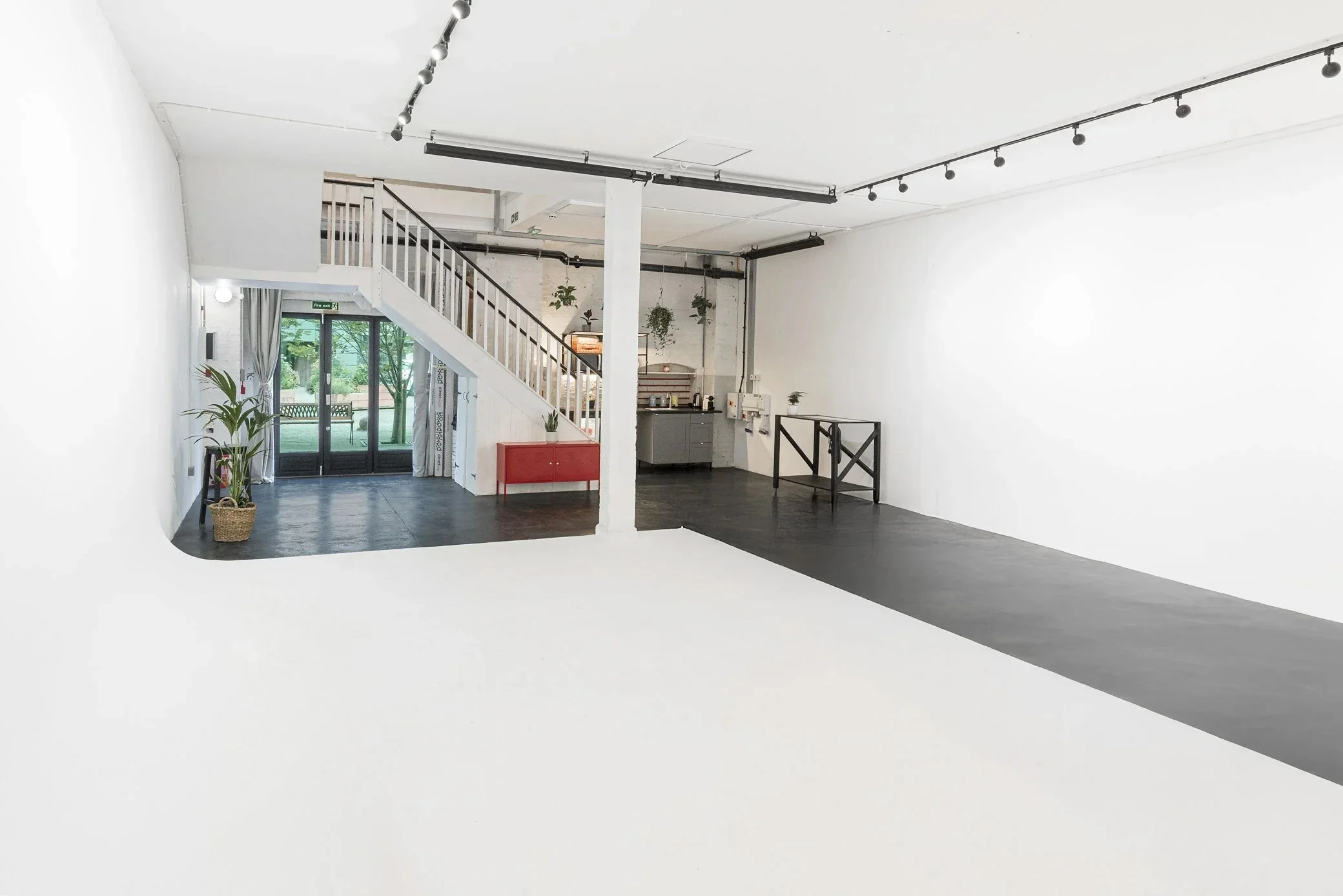 gallery cove studio space with white walls, black flooring, and a white staircase leading to a second level. There is a large glass door at the back with a view of greenery outside, and a potted plant near the entrance.