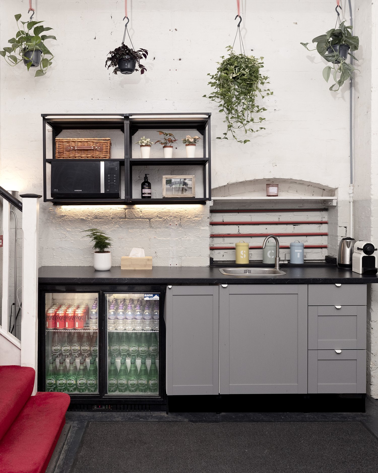 Small kitchenette with gray cabinets, a black countertop, and a microwave. Hanging plants and shelves hold decor and a basket. Below, a mini-fridge with beverages is visible. On the counter, a coffee machine, mugs, and a sink are present.