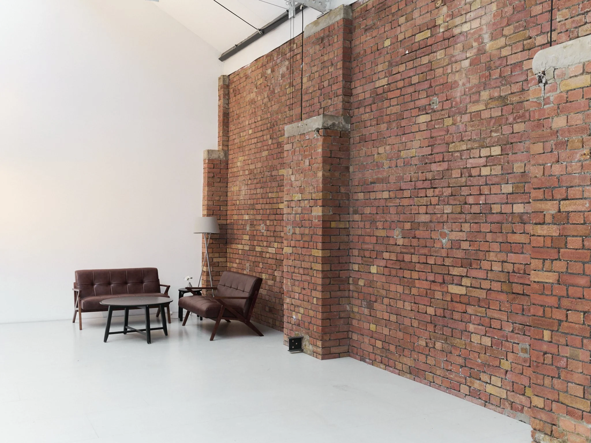 A corner of a room with a white wall on the left and a red brick wall on the right. There is a brown leather sofa, a matching bench, a round black coffee table, and a floor lamp in the corner.