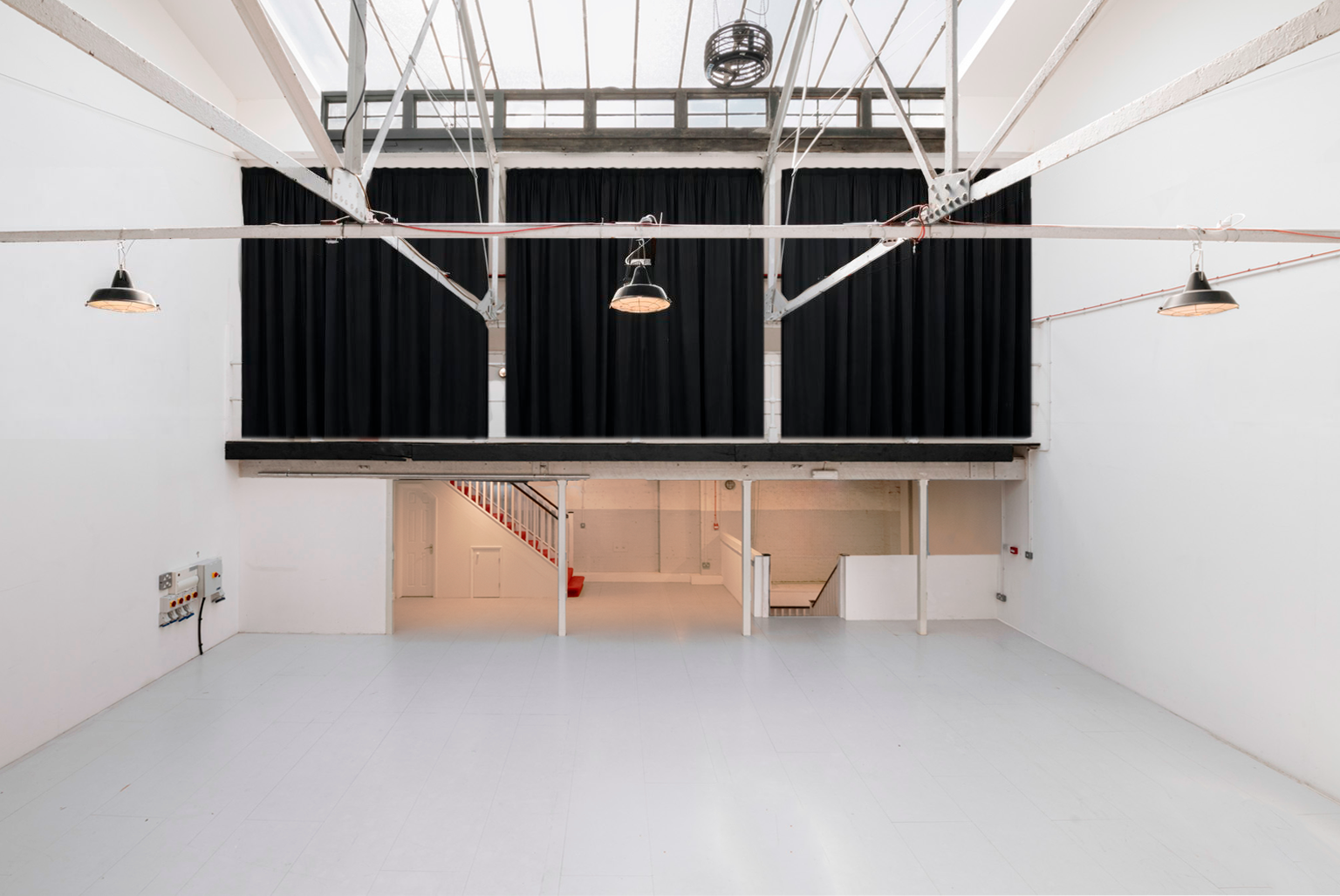 Empty spacious room with white walls, black curtains, industrial lighting, and a mezzanine level with stairs having a red handrail.