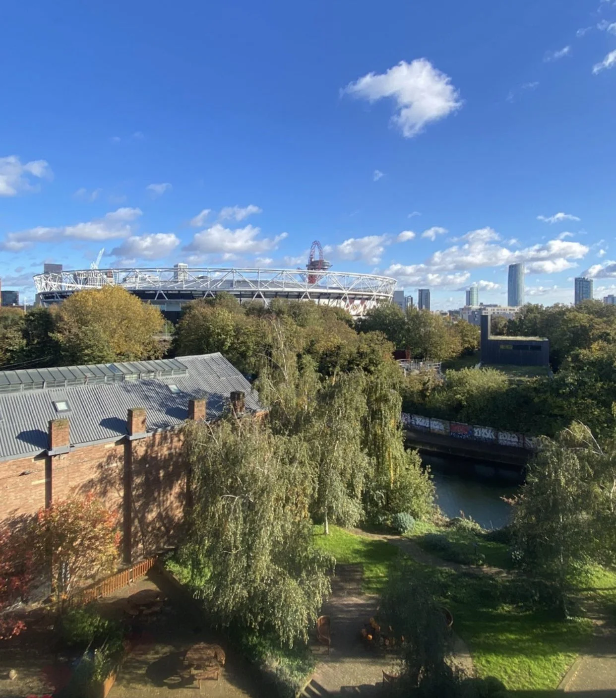 Cityscape with a sports stadium in the background, trees and a park in the foreground, a canal, and a blue sky with a few clouds.