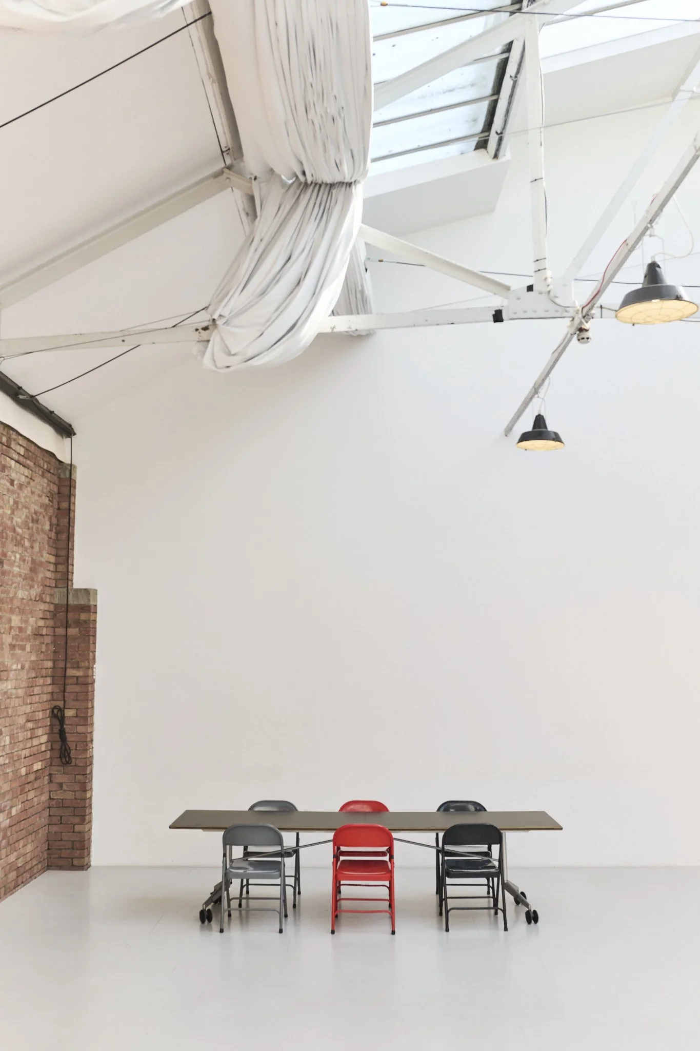 Empty room with a long table and six chairs, three black, two gray, and one red, against a white wall. Exposed brick wall on the left and a high ceiling with industrial light fixtures.