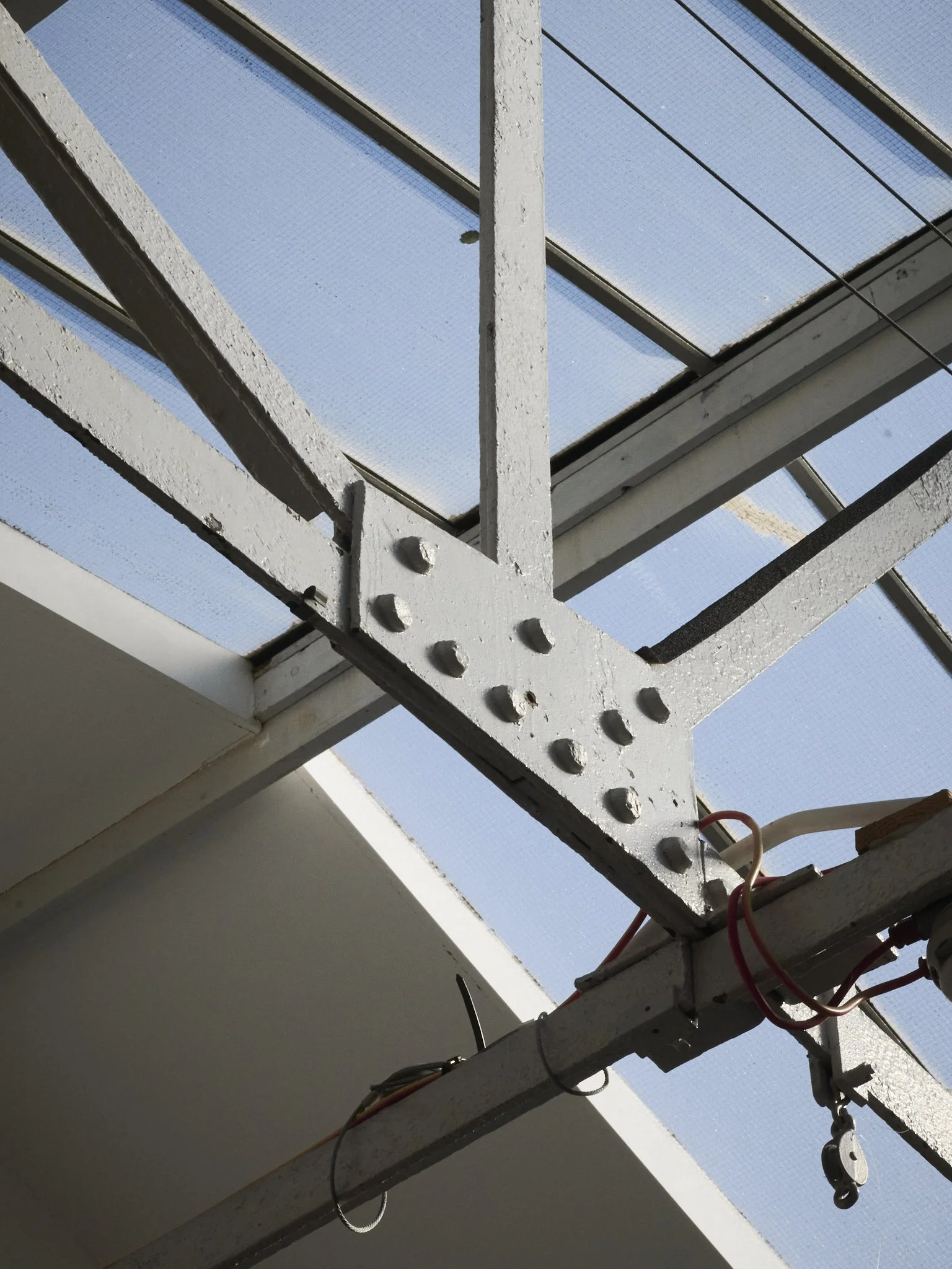 Close-up of a structural metal beam with bolts, part of a window frame or skylight, with a mesh screen and clear blue sky visible outside.