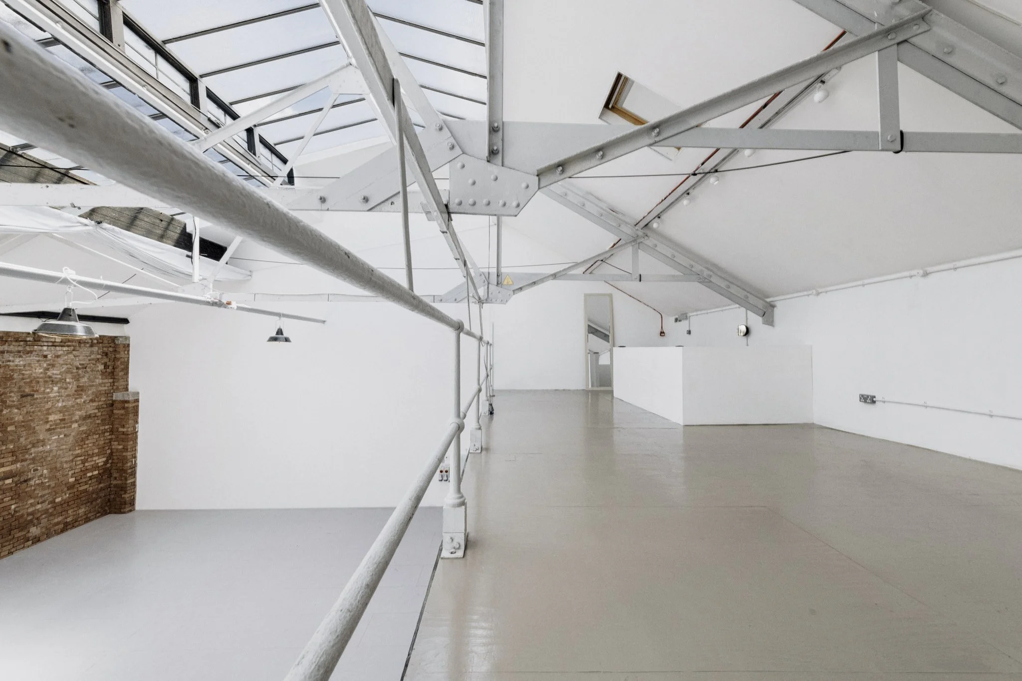 Empty industrial-style gallery or event space with white walls, high ceilings, skylights, exposed metal beams, and a brick accent wall.