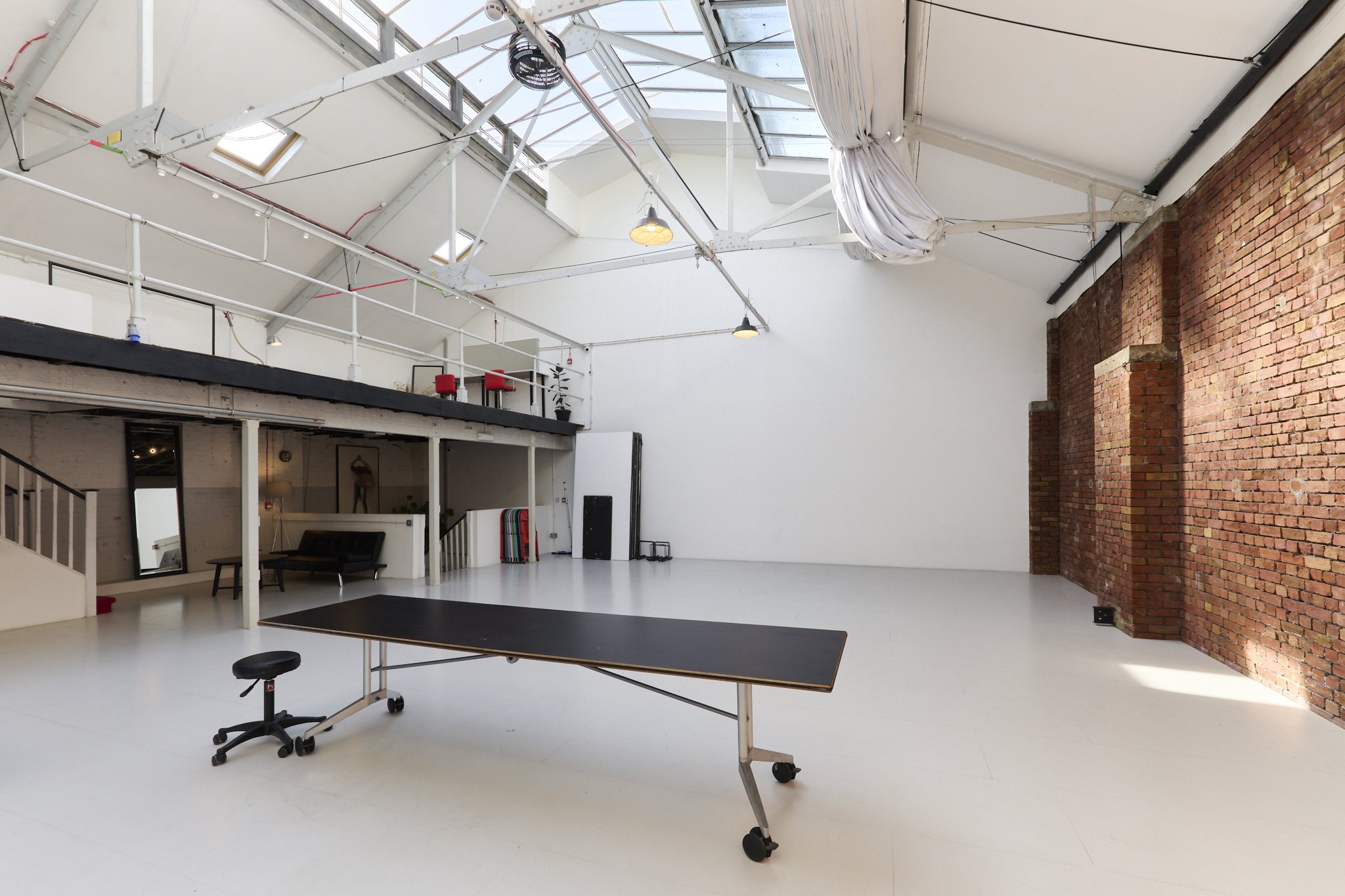 Spacious industrial loft with brick wall, high ceiling, large windows, and movable furniture including a black table on wheels and a stool.