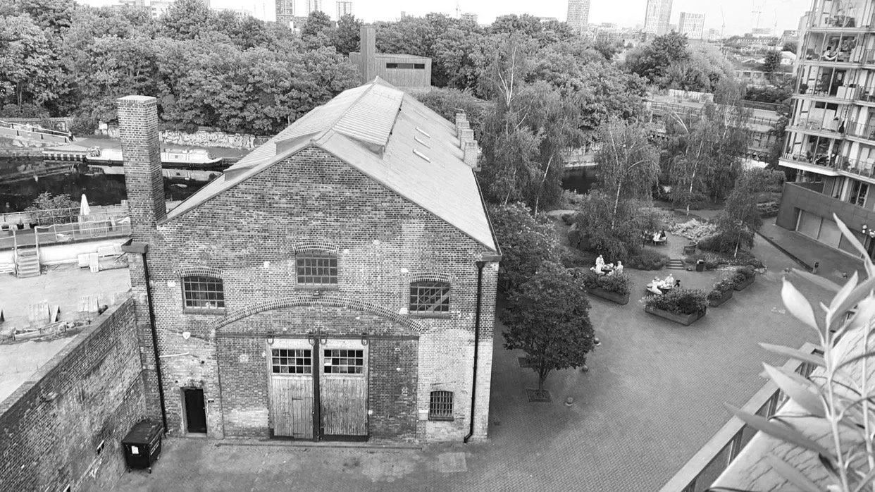 Black and white photo of an old brick building with a large garage door, windows, and a chimney, overlooking a courtyard with benches, trees, and a body of water with boats in the background, high-rise buildings in the distance.