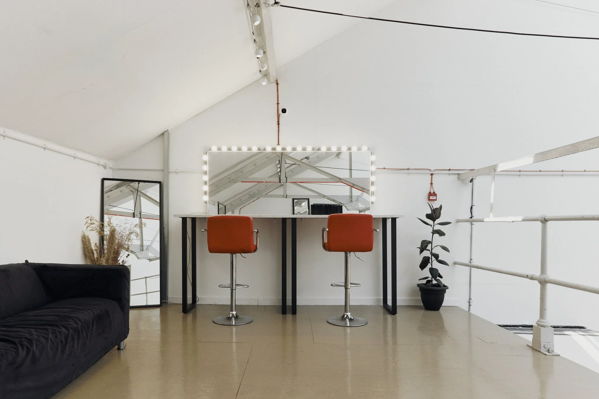 Interior of a modern loft with a black sofa, a high table with two orange chairs, a large mirror with lights, another smaller mirror, a potted plant, and white walls with exposed wiring.