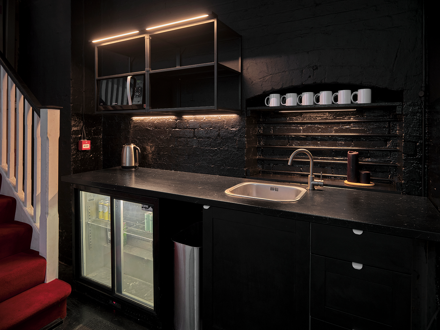 Small black kitchen area with a black countertop, stainless steel sink, and a black backsplash. Open shelving with white mugs, a microwave, and a small open shelf with candles. A mini fridge with glass door and beverages inside, a kettle, and trash b