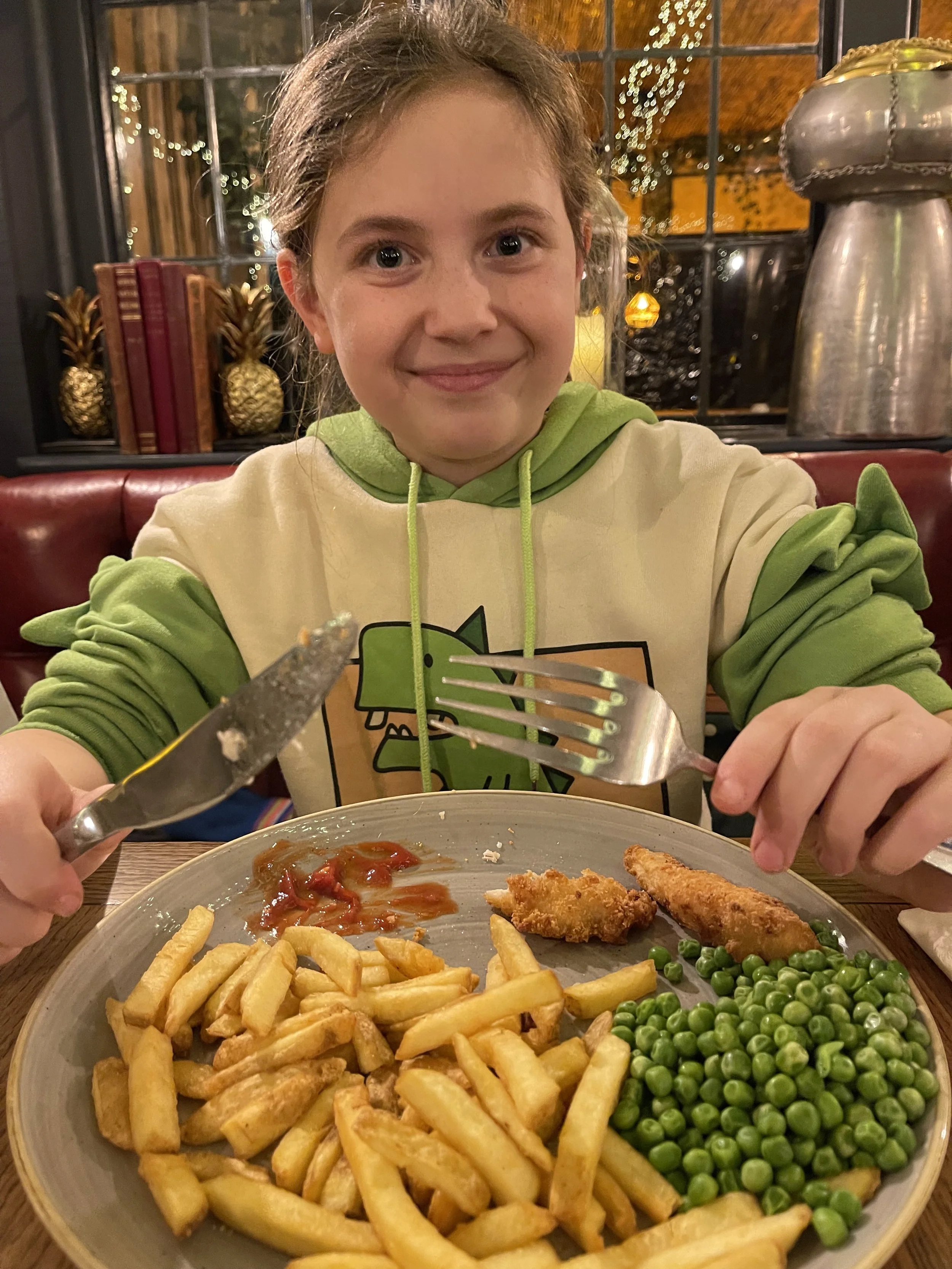 Child in a hoodie dining on French fries, chicken strips, and peas at a restaurant.