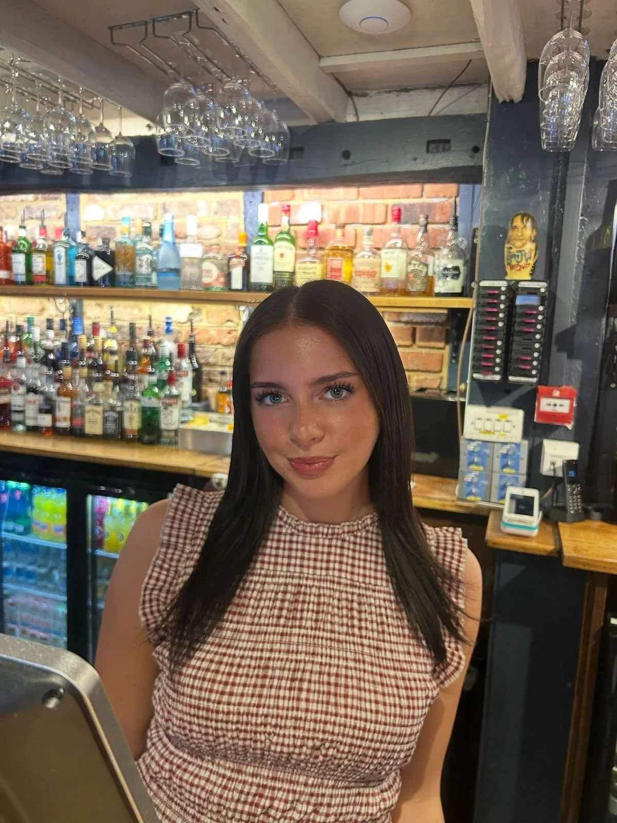 A woman with blonde hair serving draft beer from a Peroni tap in a bar. She is smiling and wearing a black shirt and striped apron, with glasses on her head. Behind her are shelves stocked with various liquor bottles and a brick wall.