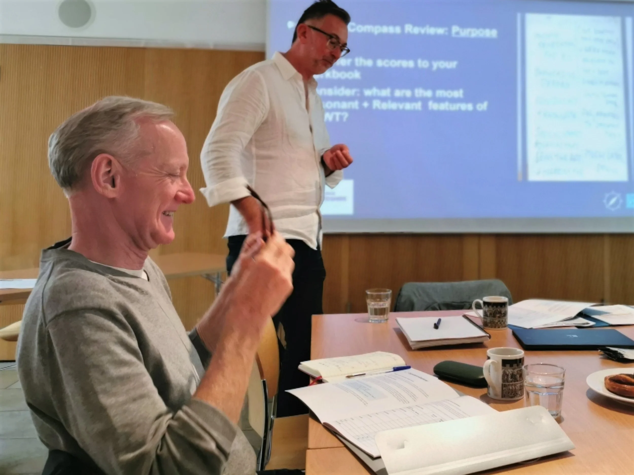 A man at a table with a man standing up next to time in front of a presentation on a screen