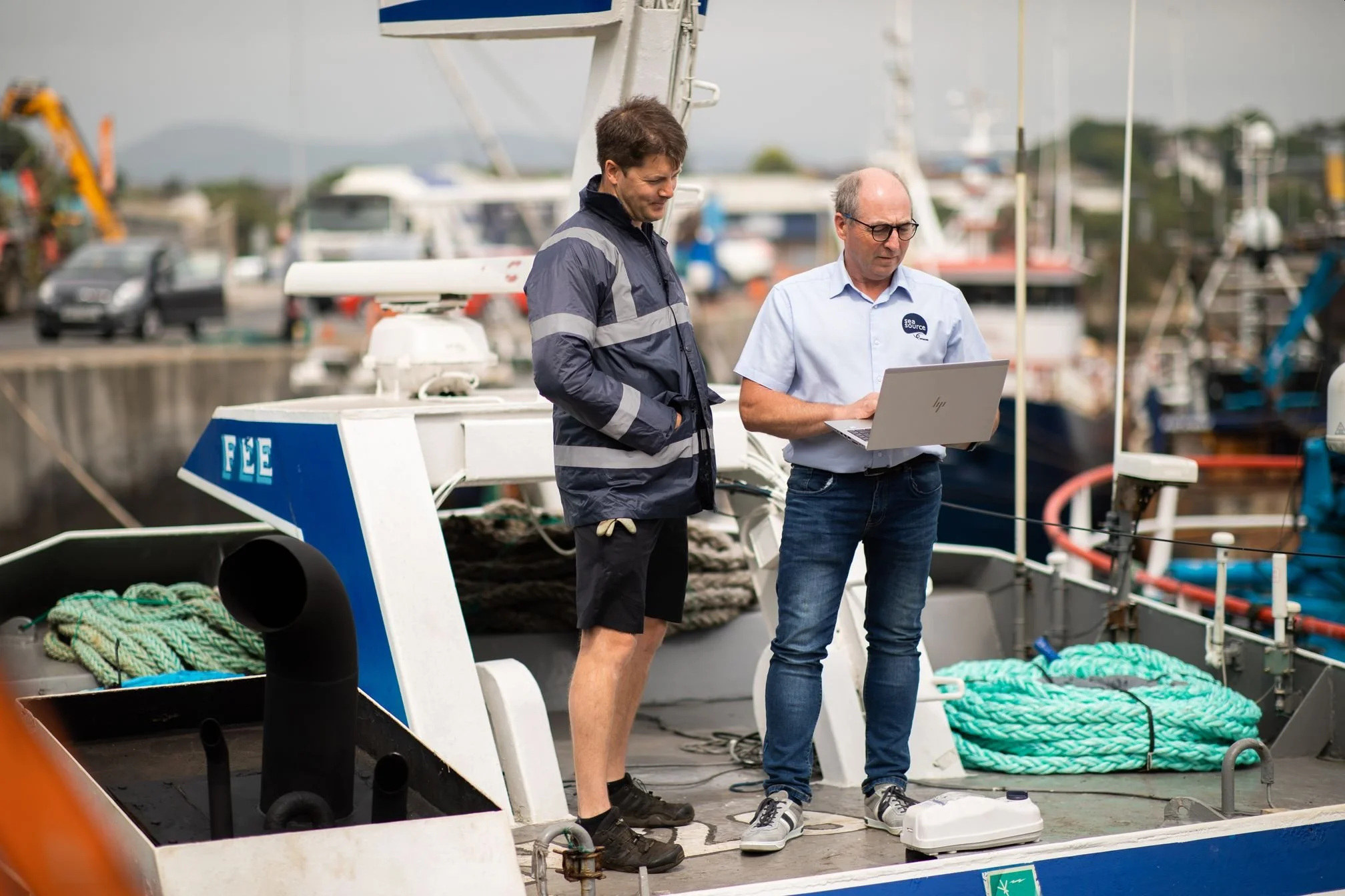 Two men standing on a boat docked at a marina, one holding a laptop while the other looks at the screen, with boats and vehicles in the background.