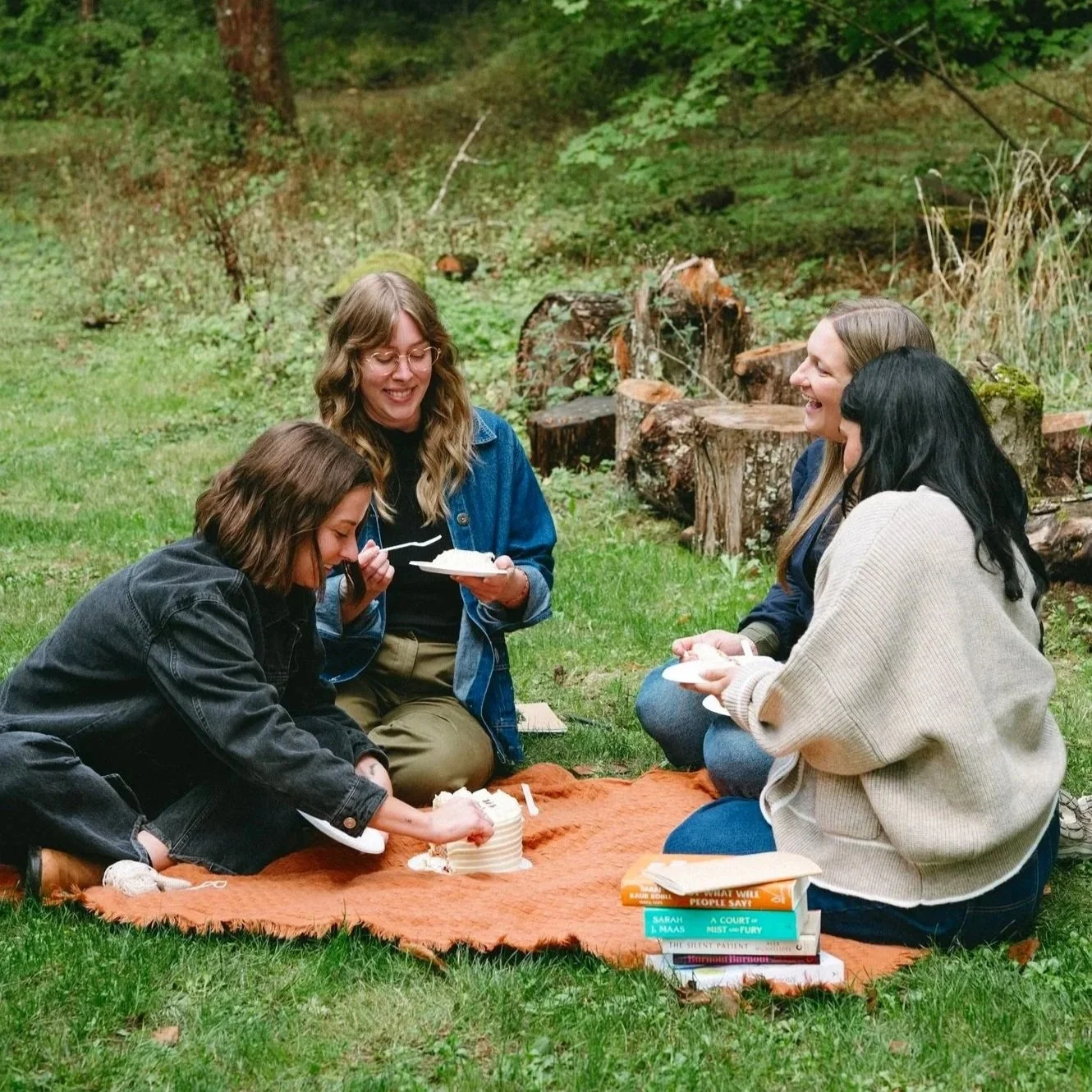 Four women sitting outdoors on a blanket, celebrating a birthday with a small cake, surrounded by trees and logs.
