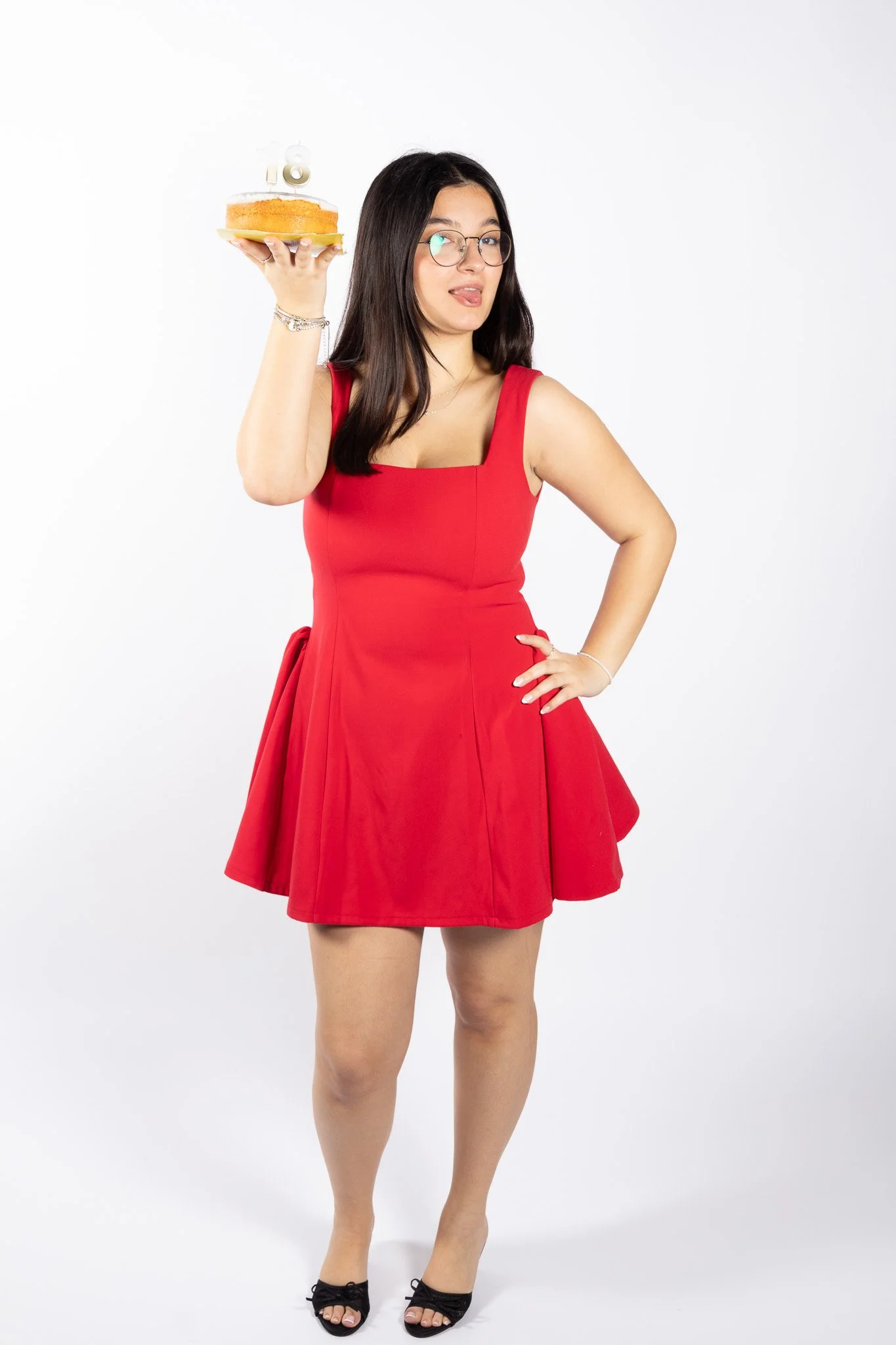 A young woman in a red dress holding a birthday cake with lit candles, sticking her tongue out, against a plain white background.
