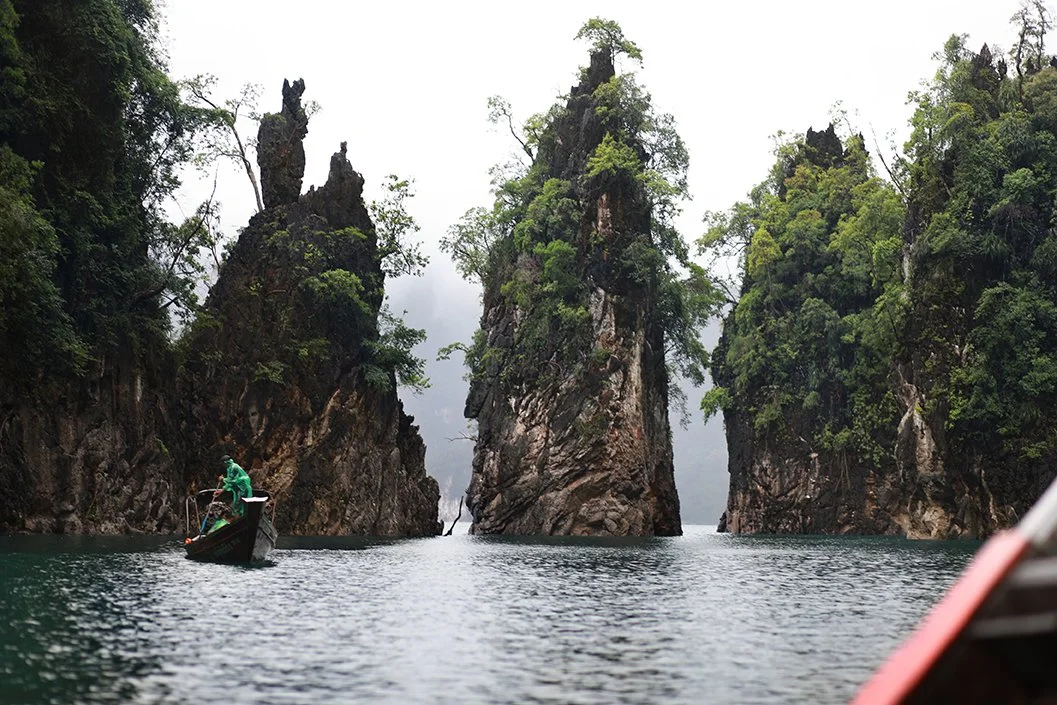 A boat with a person in green clothing navigating through a narrow waterway between tall, green-covered rocky islands under a cloudy sky.