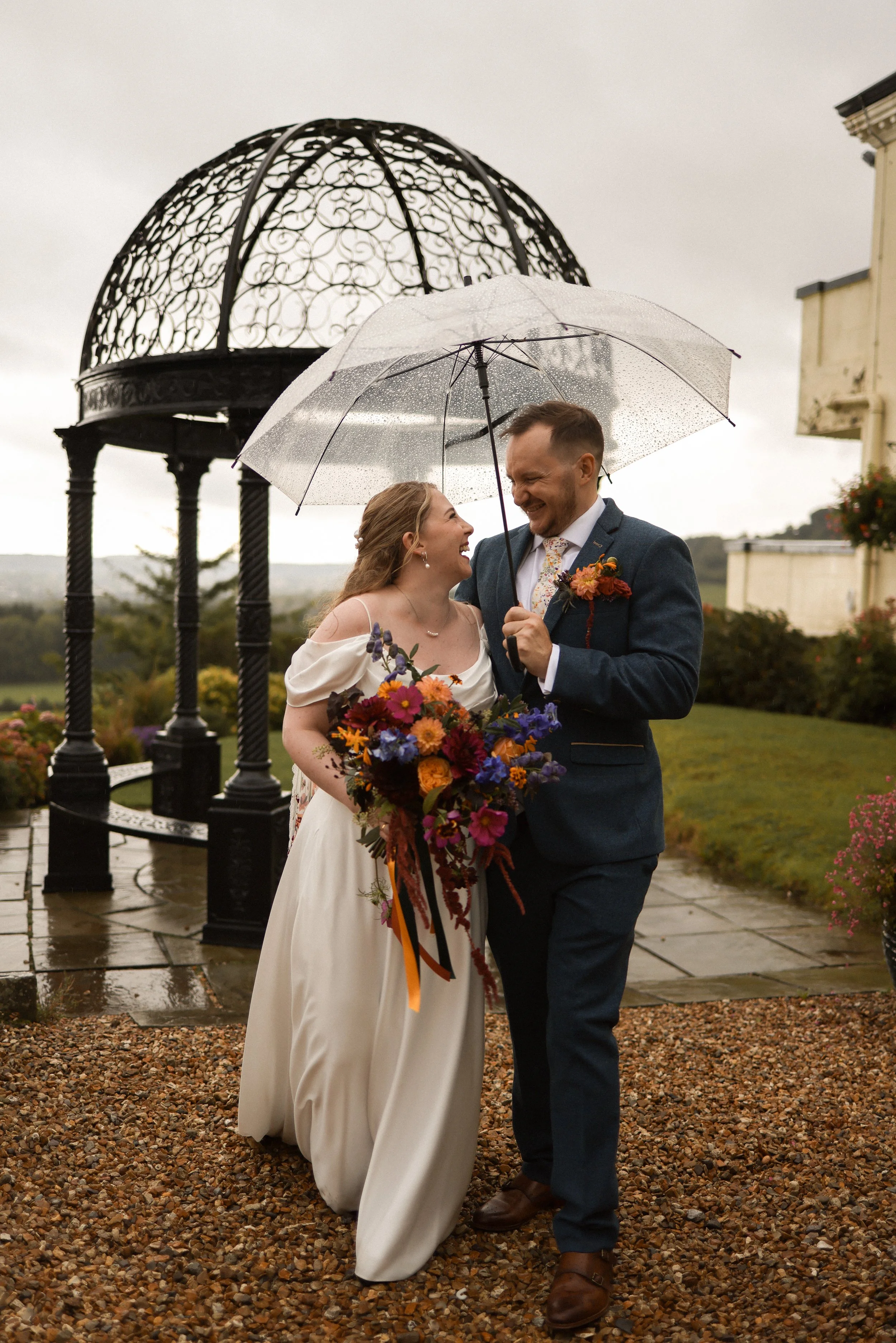 The happy couple stood under an umbrella in the rain holding, smiling at each other, with the bride holding her jewel-toned bridal bouquet with coloured ribbon