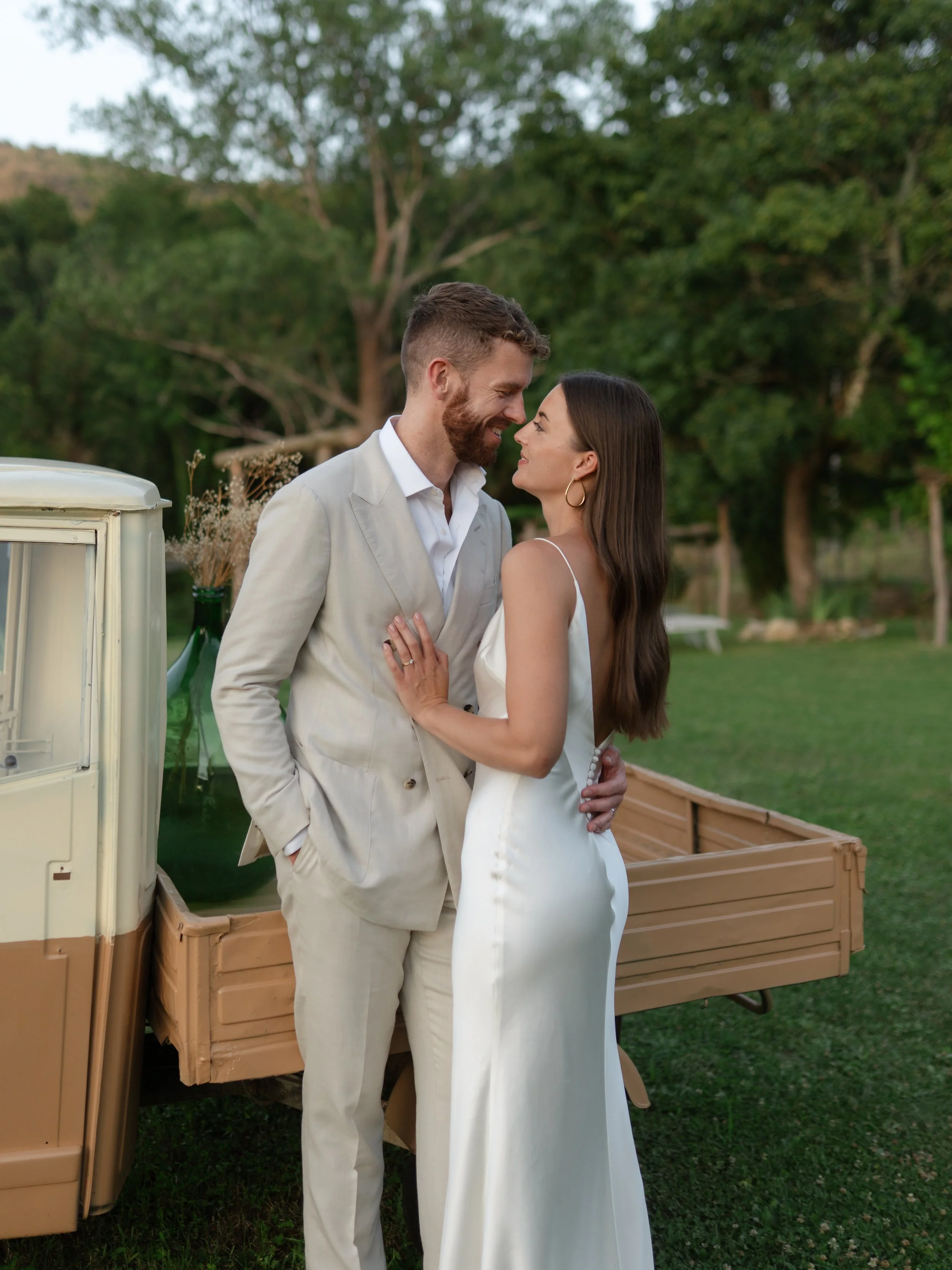 A couple in wedding attire holding hands and walking outdoors near a rustic building, with a hill covered in greenery in the background, during sunset.