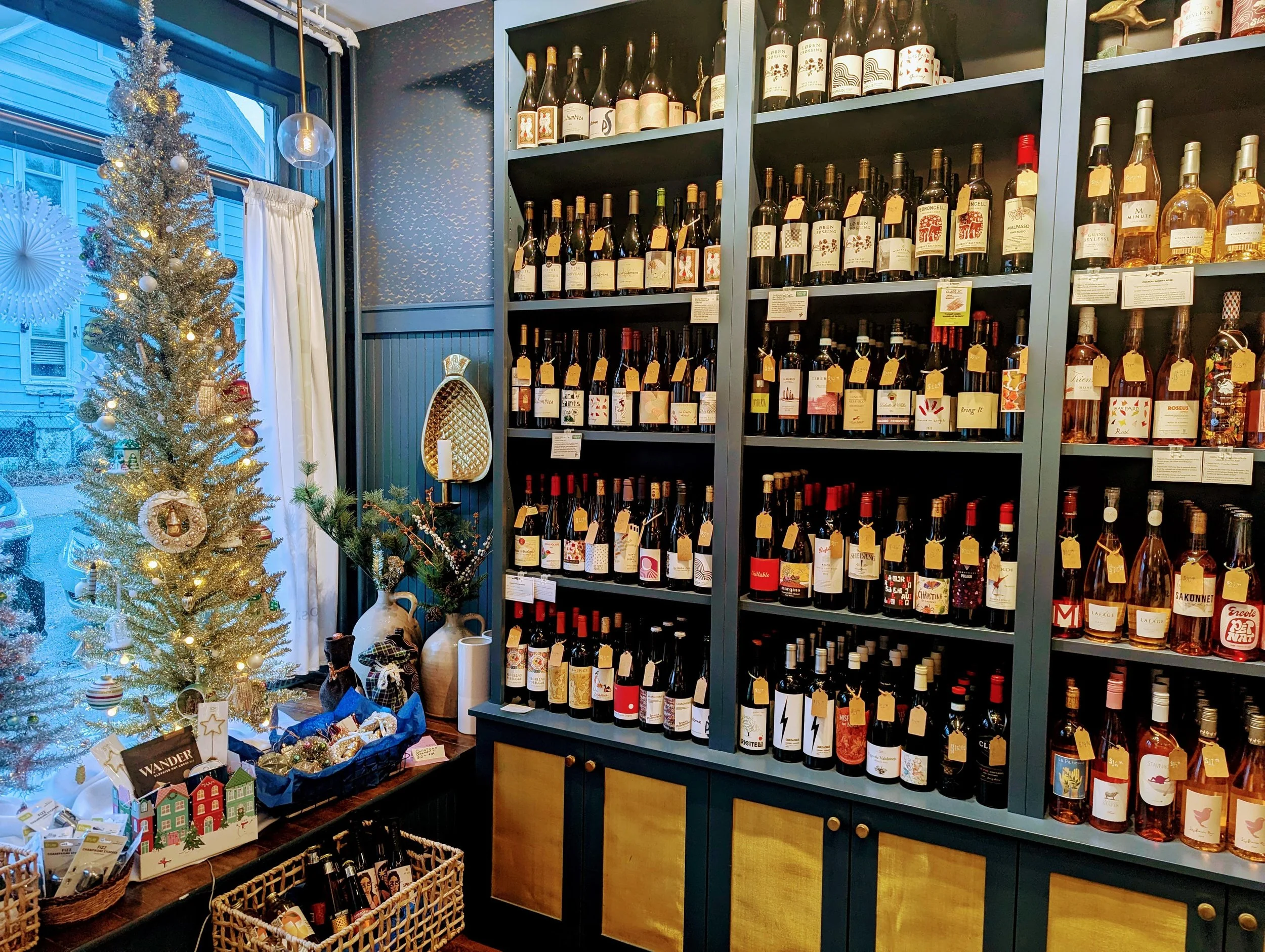 A cozy indoor space with a Christmas tree decorated with ornaments and lights in front of a window. To the right, there are large shelves filled with bottles of wine and liquor, each with a small yellow tag. In front of the shelves, there are baskets