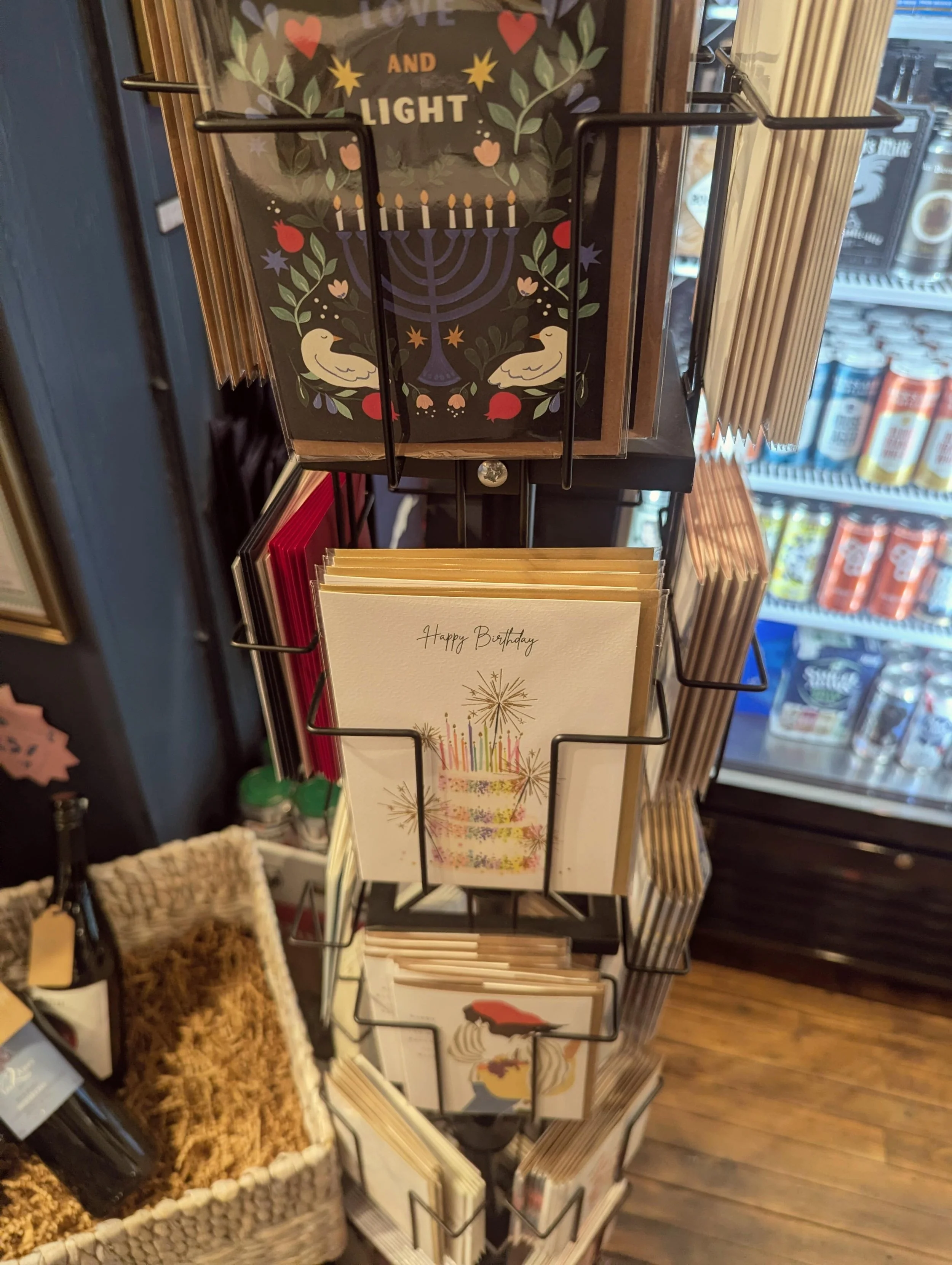 A display stand with greeting cards, including a birthday card with a cake illustration and fireworks, and holiday cards with festive designs, in a store with a refrigerator in the background.