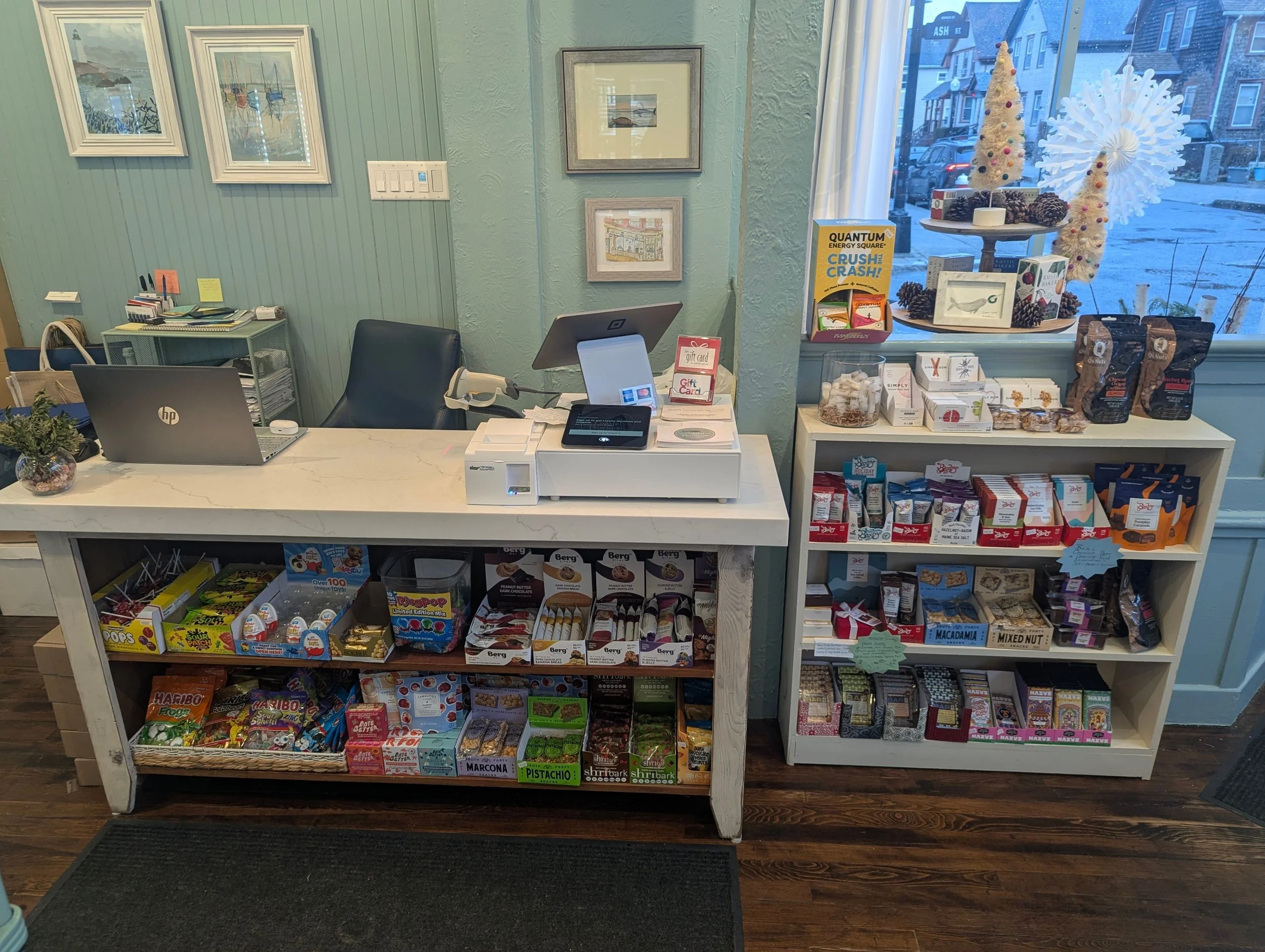 A cozy retail checkout counter with a white marble top, behind which is a black chair, a laptop, and a cash register. To the right, a white shelf displays an assortment of packaged snacks and chocolates, with holiday-themed decorations on a nearby wi