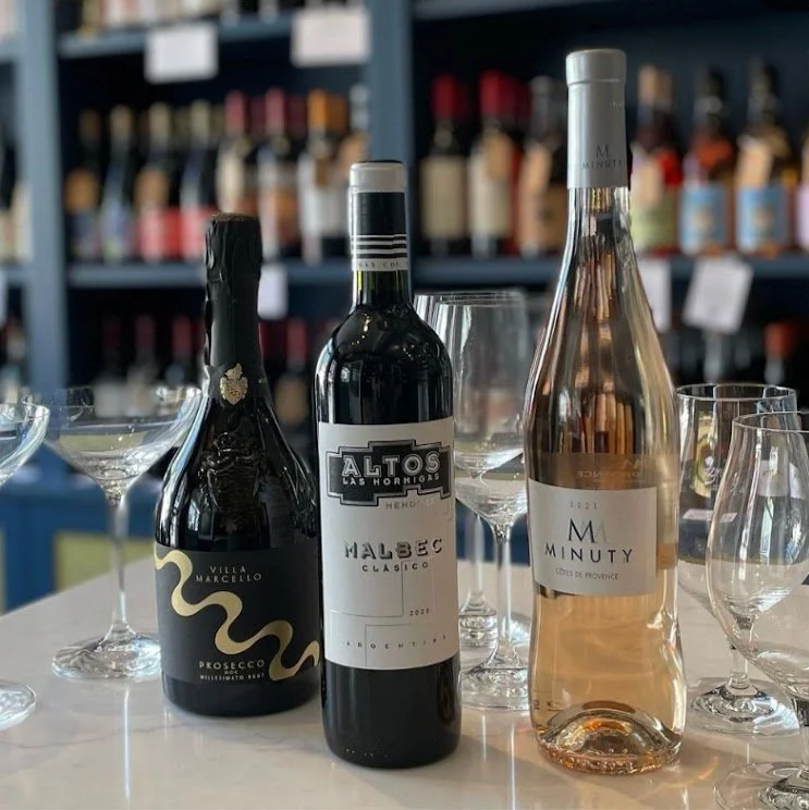 Three bottles of wine and several empty wine glasses on a white table, set in a wine shop or tasting room with shelves of wine bottles in the background.