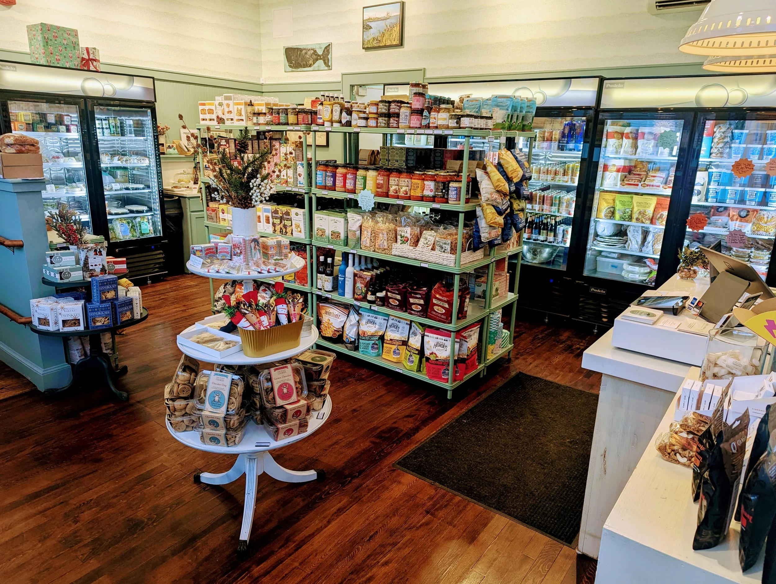Interior of a small grocery or specialty shop with shelves stocked with snacks, baked goods, and beverages, including refrigerated sections and a cash register.