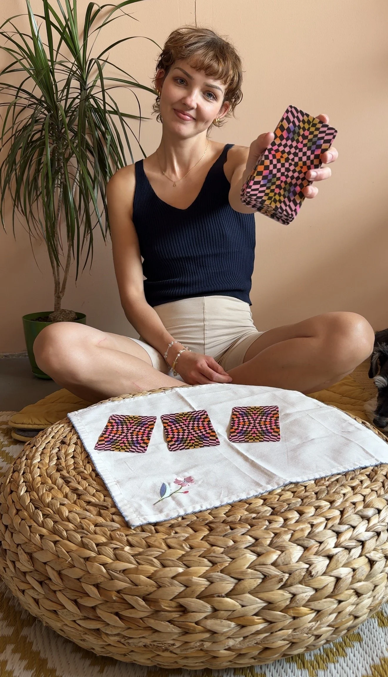 A woman with short curly hair, wearing a dark sleeveless top and beige shorts, sits cross-legged on a round woven rattan table, holding a card with a colorful checkered pattern, with three similar cards laid out on a white cloth. Behind her is a larg
