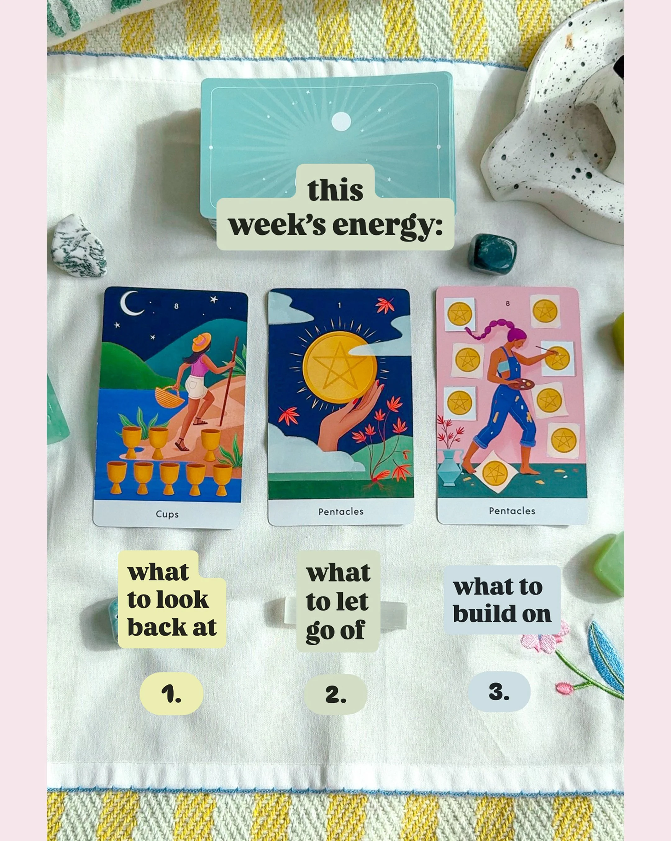 Three tarot cards are laid out on a white cloth with some stones and a vase nearby. The cards are labeled 'Cups,' 'Pentacles,' and 'Pentacles.' There are sticky notes underneath with texts: 'what to look back at,' 'what to let go of,' and 'what to bu