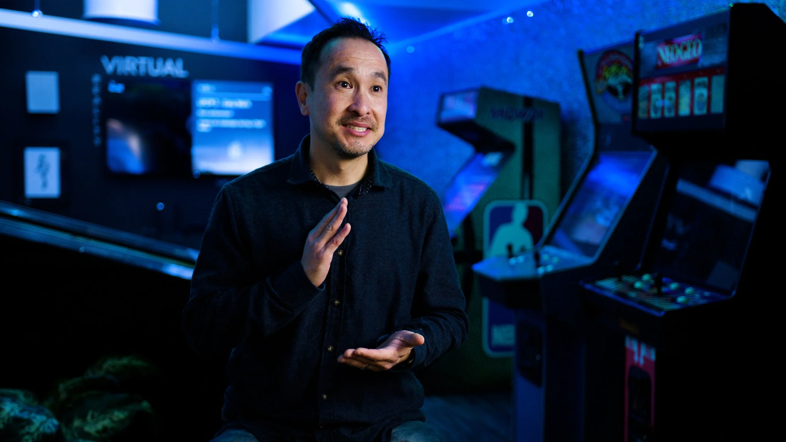 Man seated in a neon-lit gaming studio surrounded by arcade machines.