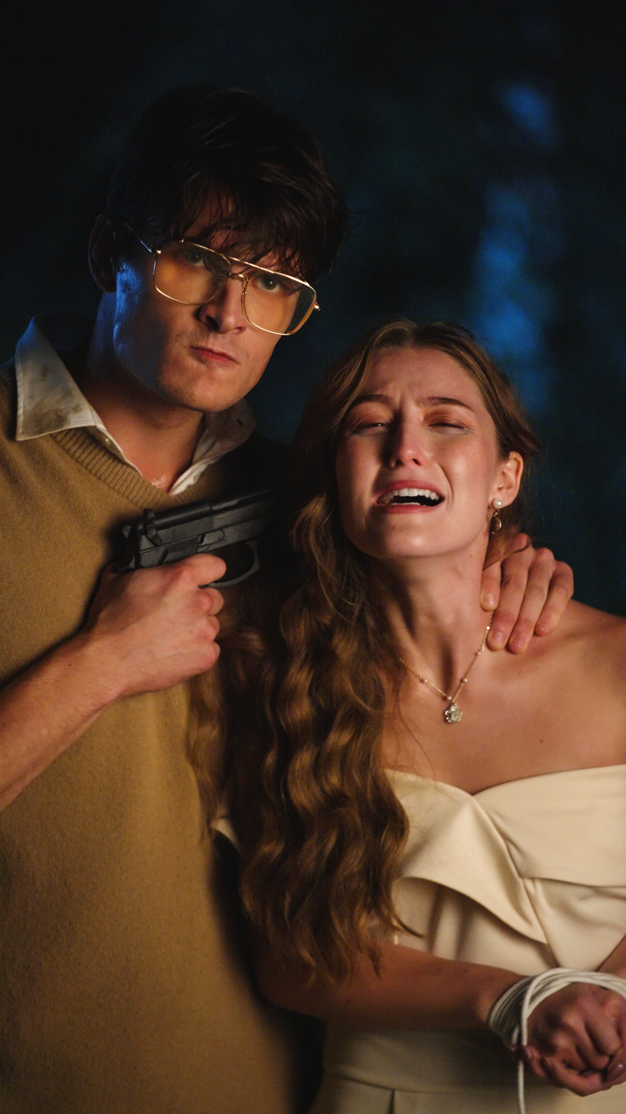 Man wearing glasses and a tan sweater vest holding a handgun against a woman in an off-shoulder white dress with her hands bound outdoors at night.