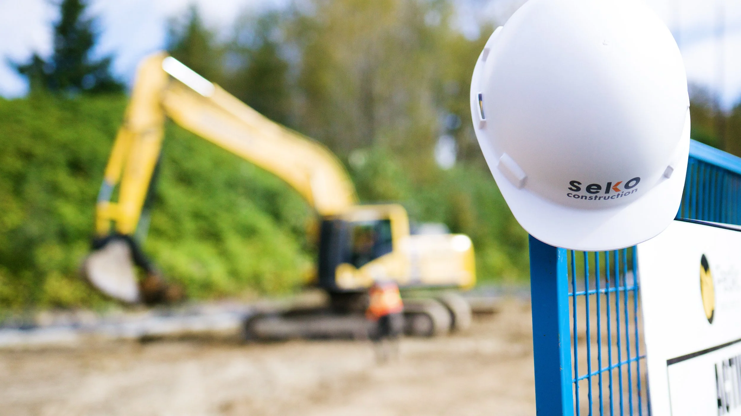 Seko Construction hard hat hanging on site fence with excavator working in the background.
