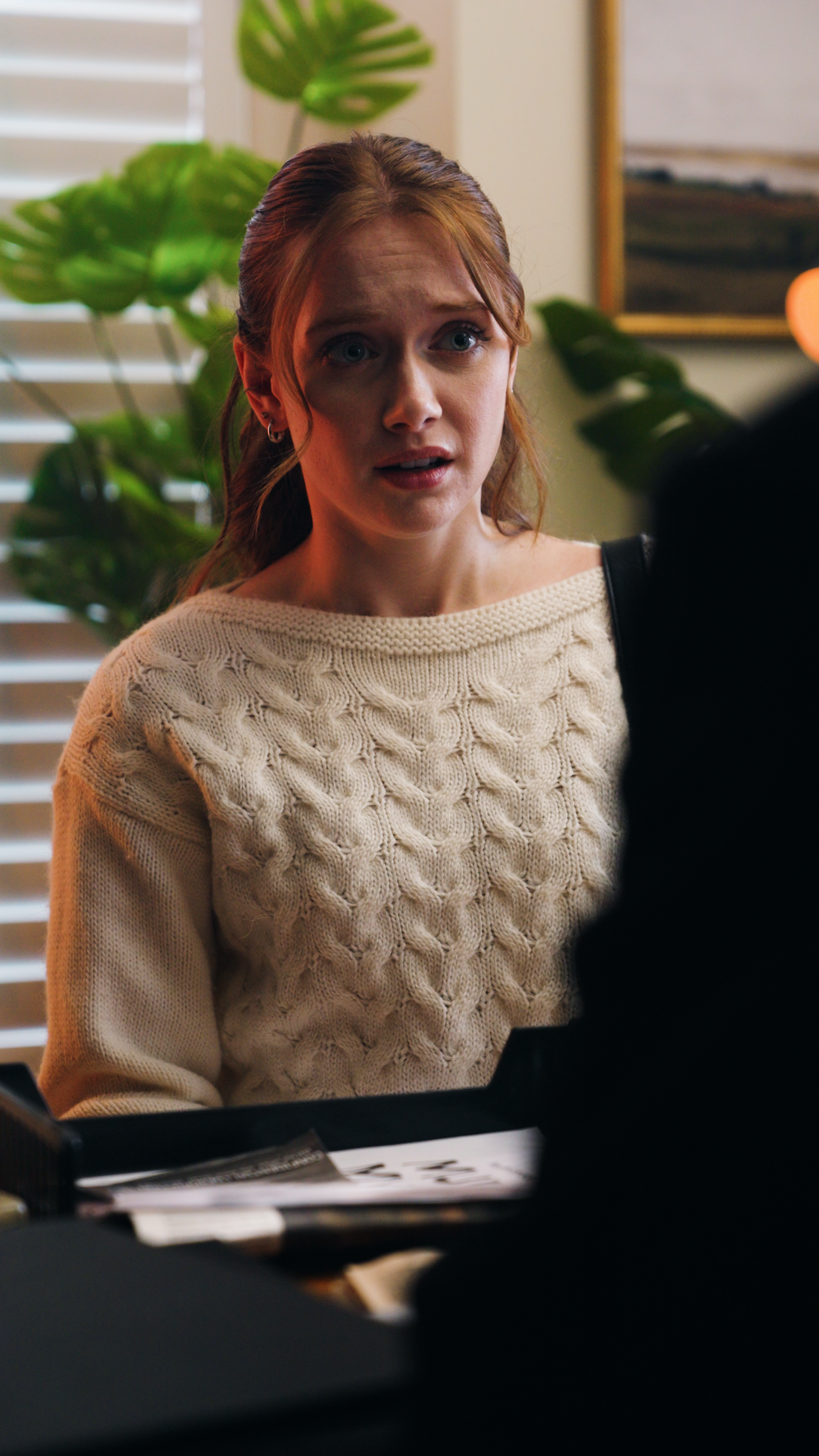 Woman in a cream cable-knit sweater seated at a desk with papers in front of her in a home interior.