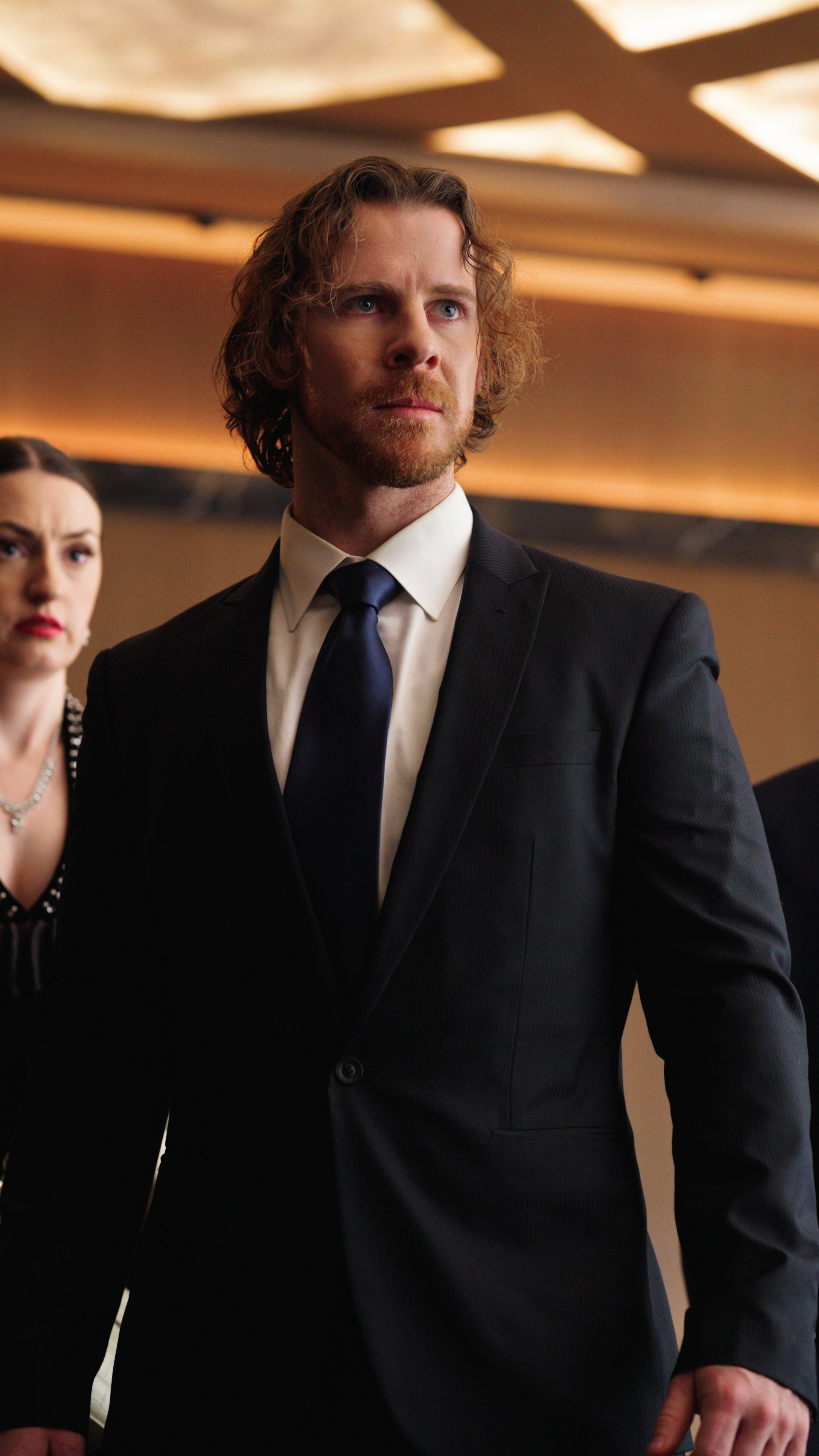 A man in a dark suit and tie stands in a modern interior with warm ceiling lighting, looking off to the side with a firm, determined expression. A woman stands slightly behind him, also dressed formally, her expression tense as they both face somethi