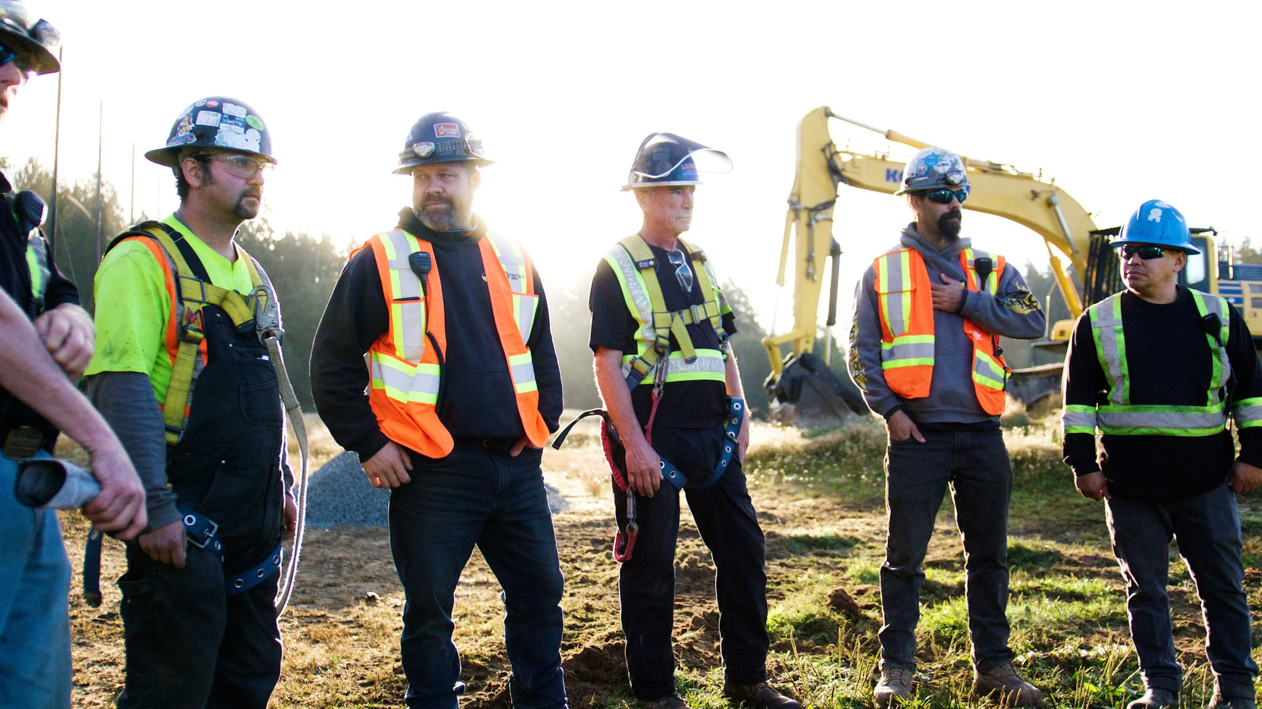 Construction crew standing together at an outdoor job site with heavy equipment in the background.