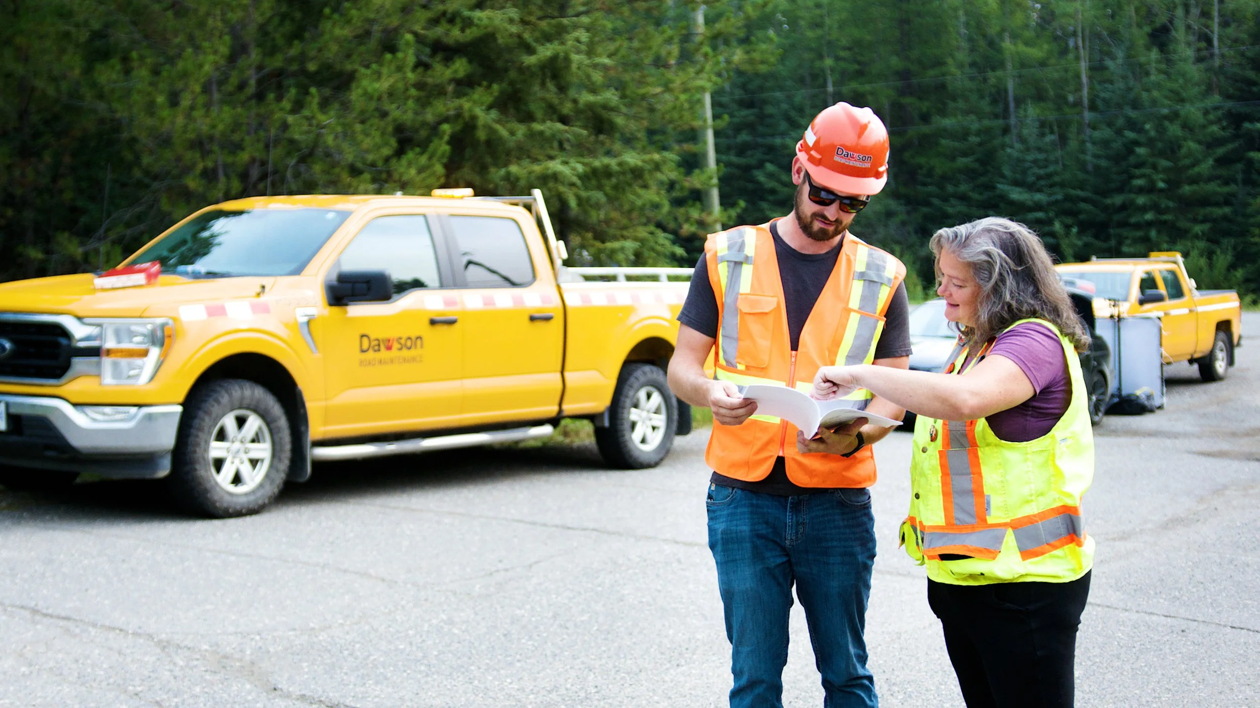 Road maintenance workers reviewing documents beside service trucks on a rural roadway.