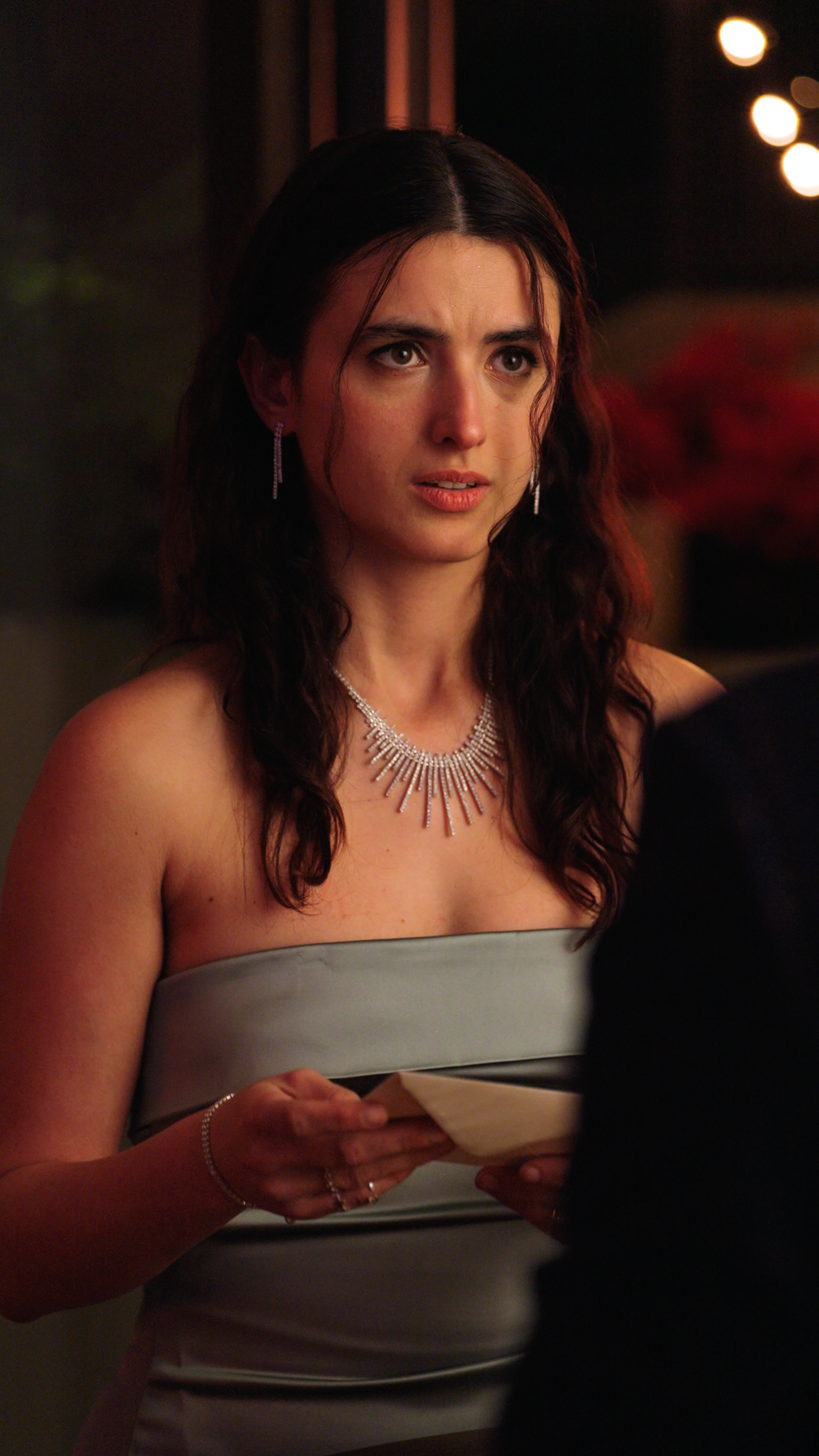 Woman in a strapless silver dress holding a folded note while standing at a candlelit dinner setting.