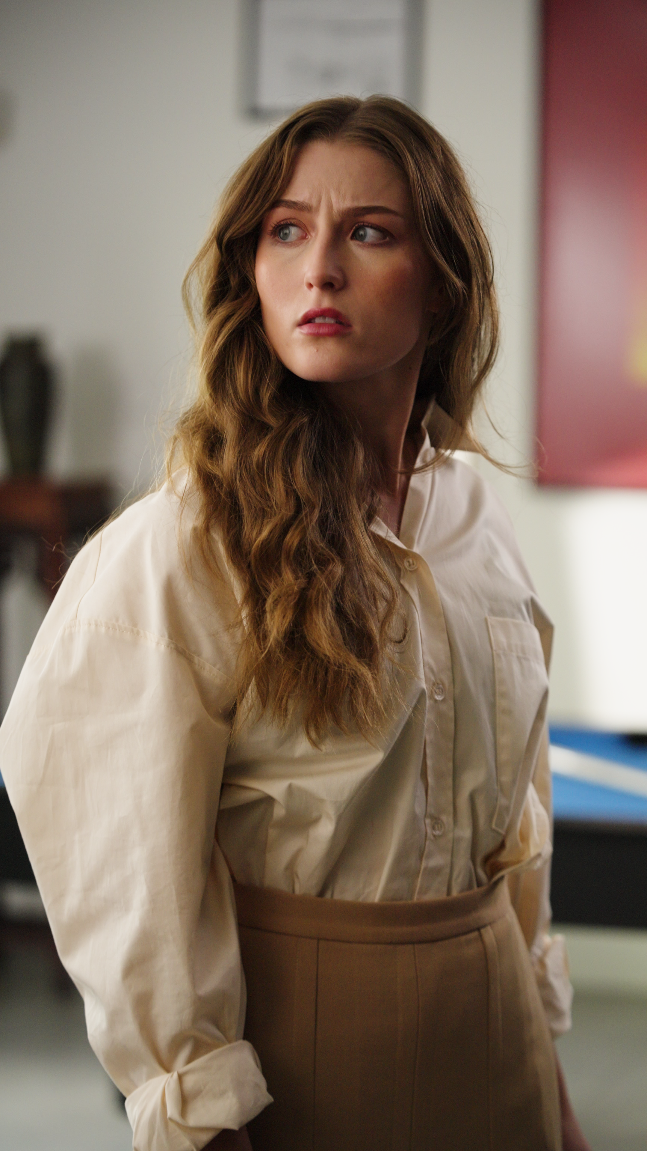 Young woman wearing a beige blouse and brown skirt looking off to the side in an interior setting.
